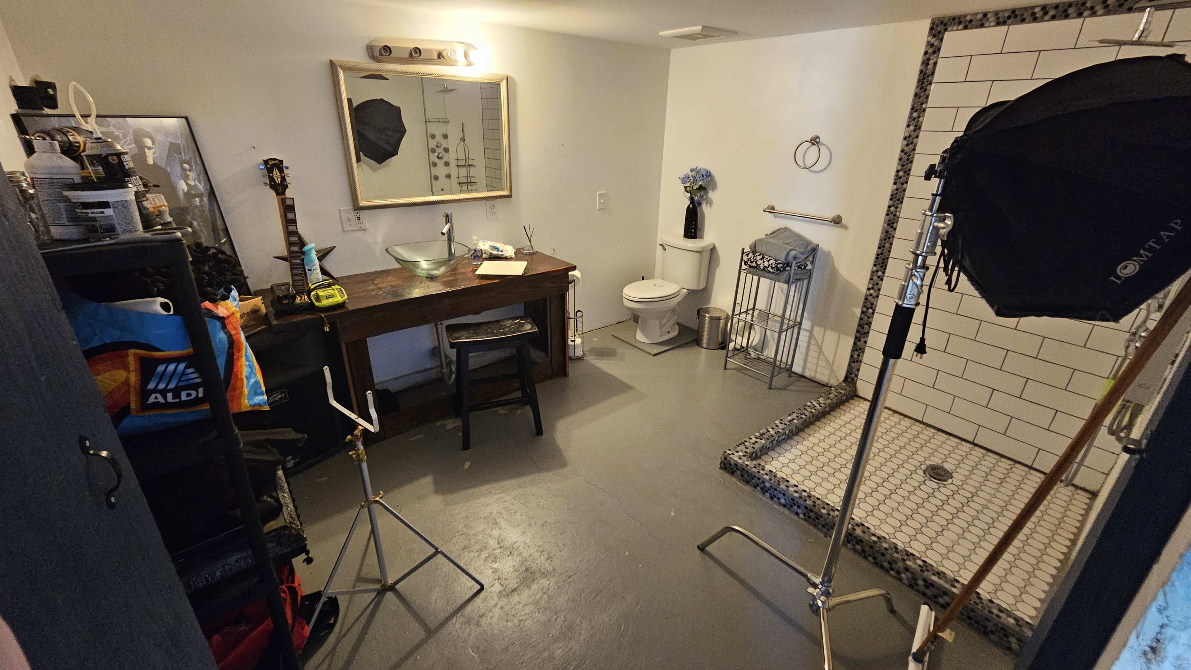 The image shows a small room with a shower area, a wooden table, a toilet, and various equipment including a light stand and props.