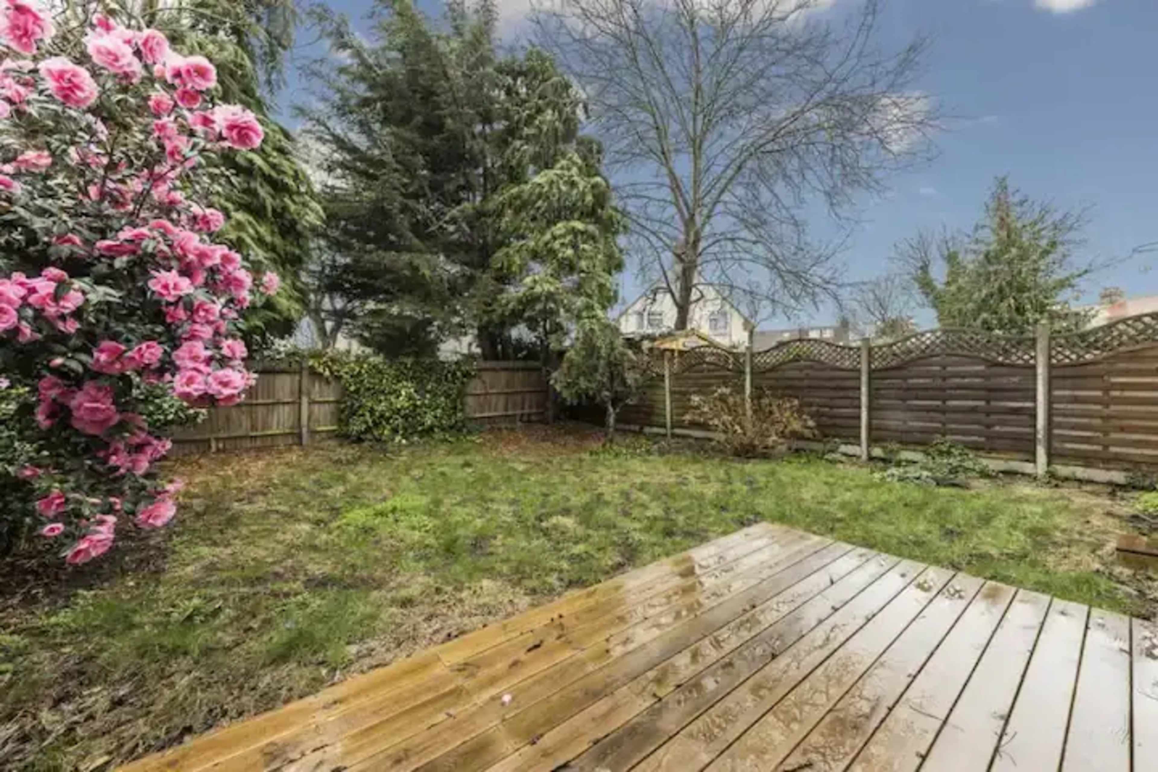 A well-maintained backyard features a wooden deck, green grass, and a flowering bush beside a wooden fence.