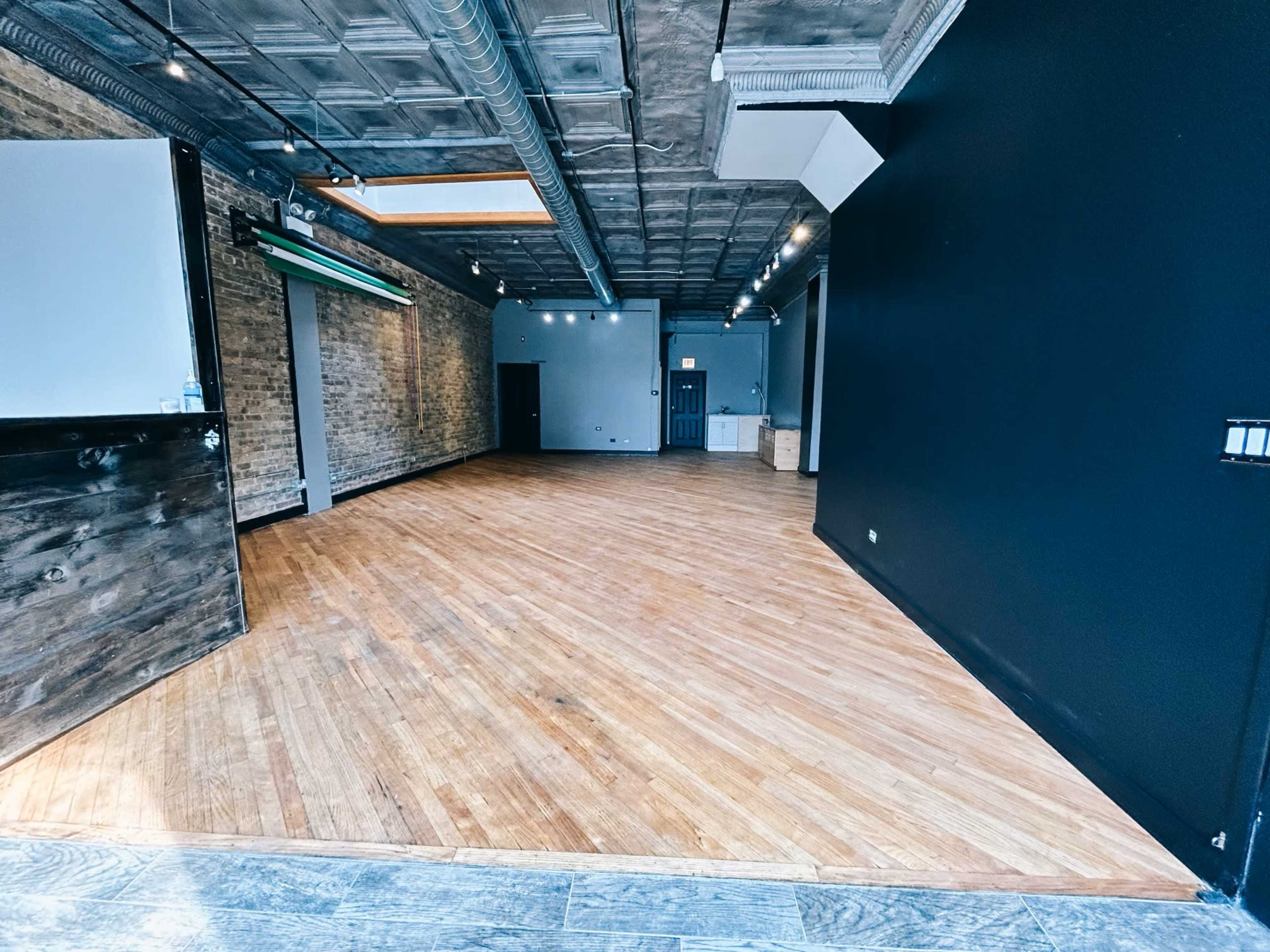 The image shows a spacious, empty interior with wooden flooring, exposed brick walls, and overhead lighting.