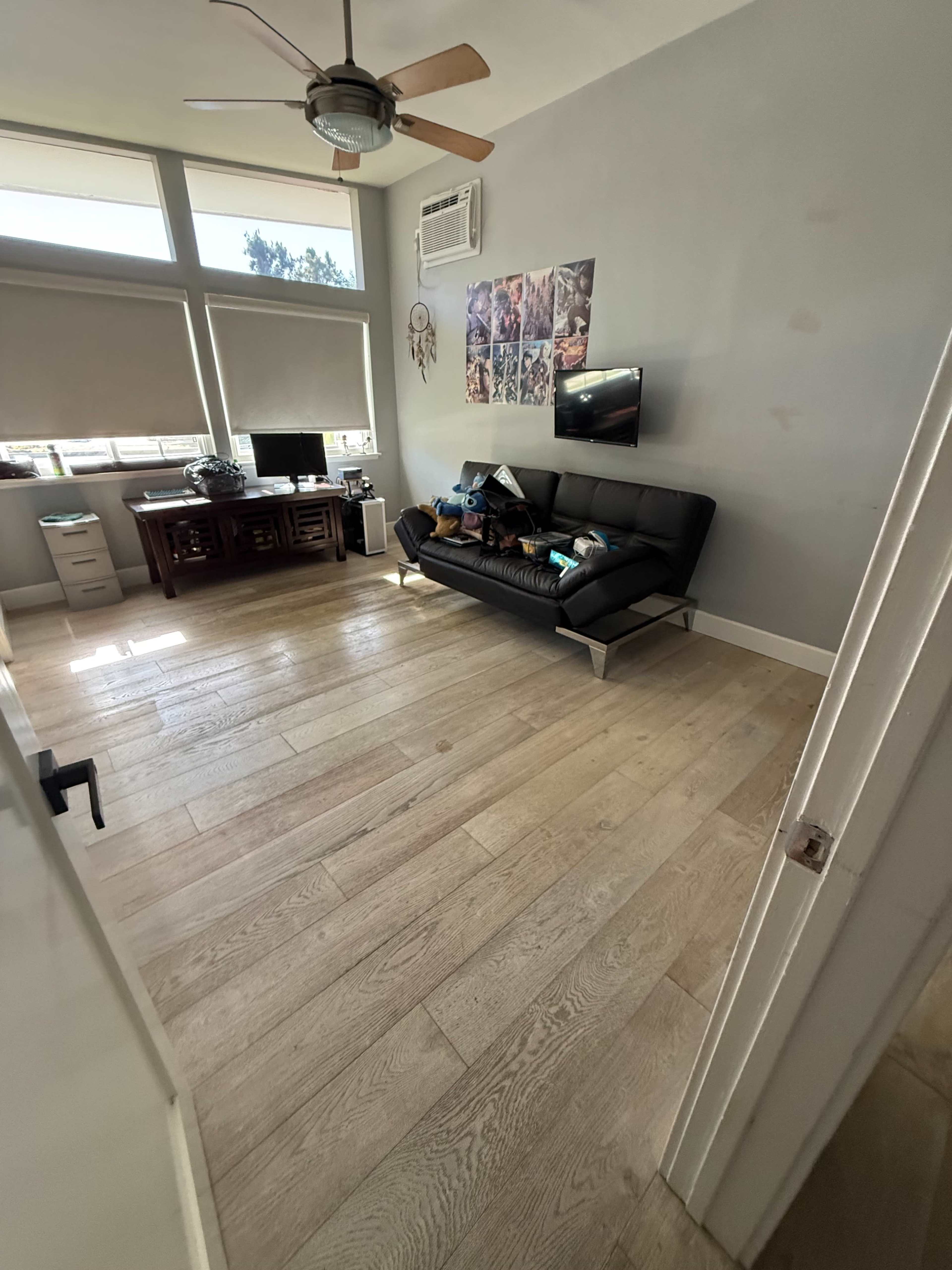 The image shows a minimalist room with a wooden floor, a black leather sofa, a desk with monitors, and various posters on the wall.