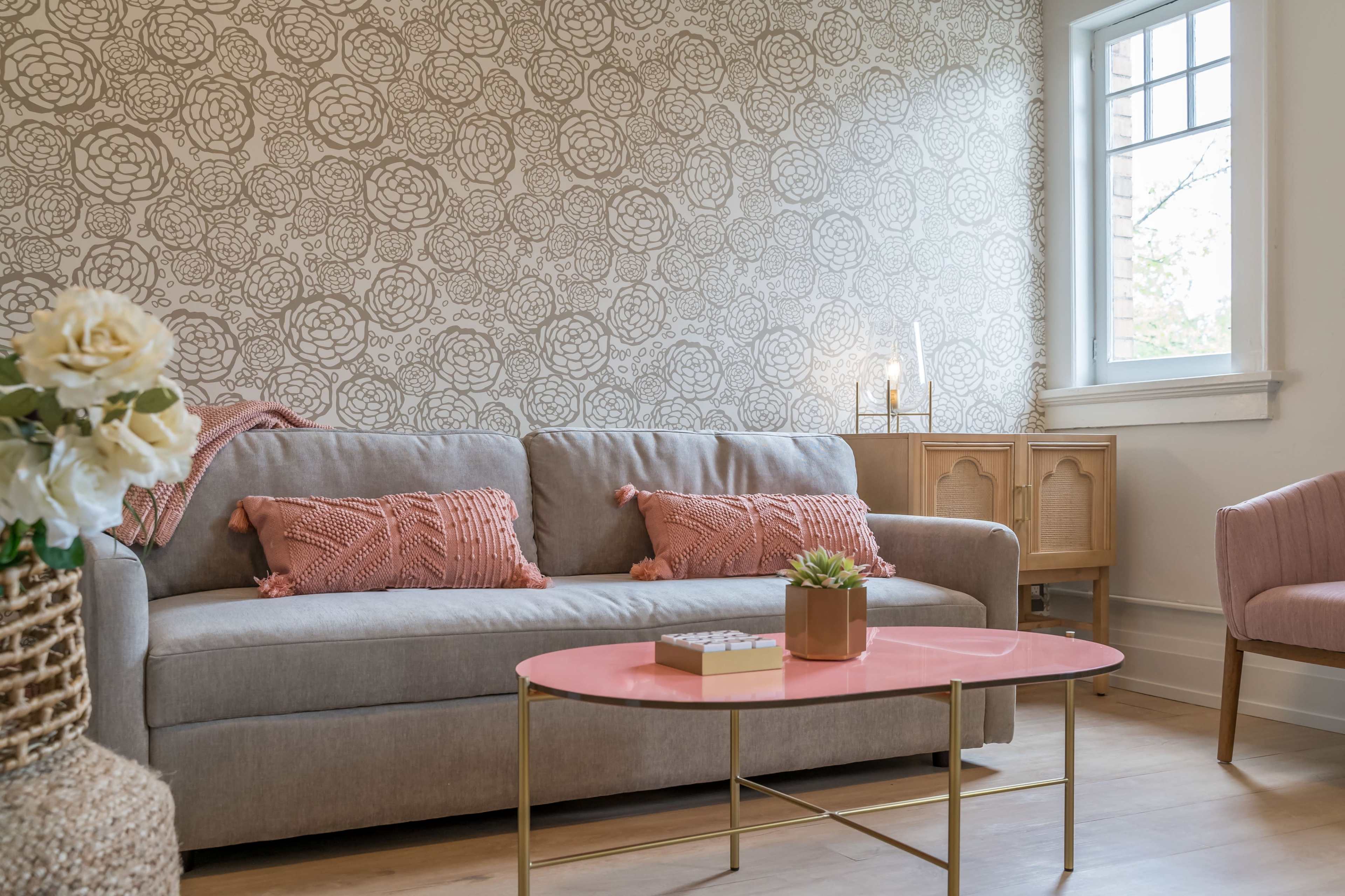 A light gray couch with pink decorative pillows is positioned in a living room featuring floral-patterned wallpaper and a pink coffee table.