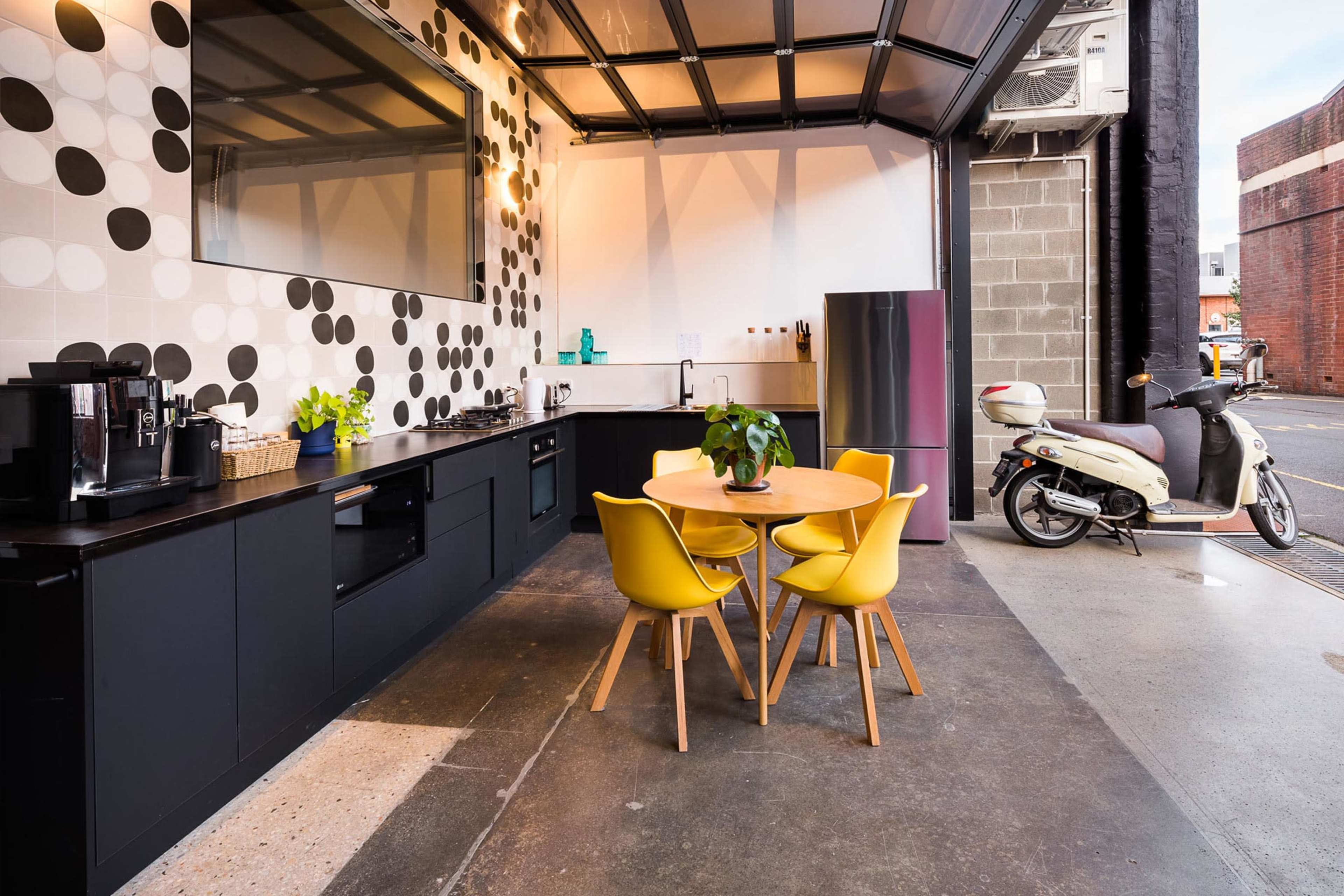 A modern kitchen with a circular dining table and yellow chairs, featuring patterned wall tiles and a motorcycle parked outside.