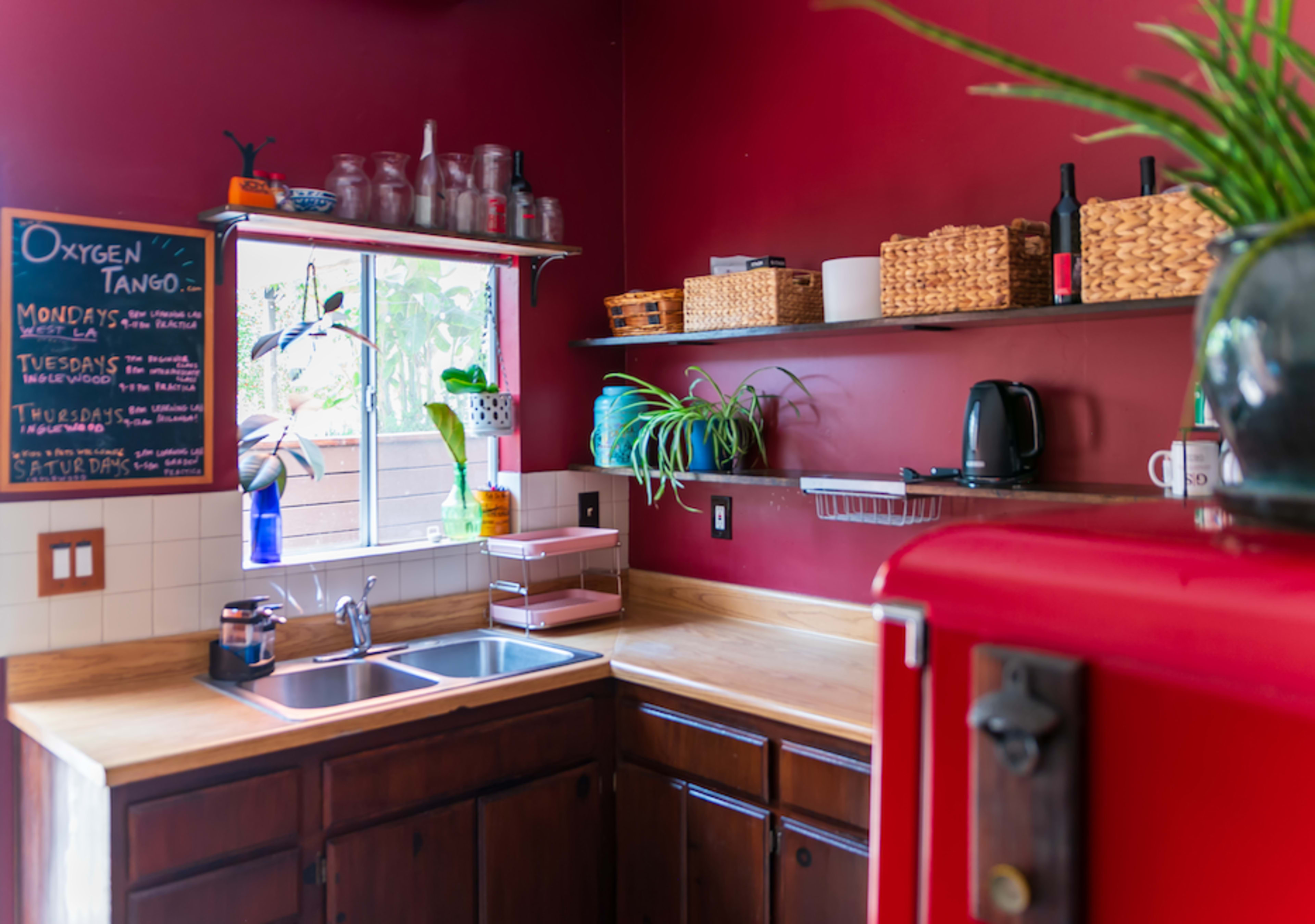 The kitchen features dark wood cabinets, a double sink, open shelving with various containers, and a red refrigerator against a deep red wall.