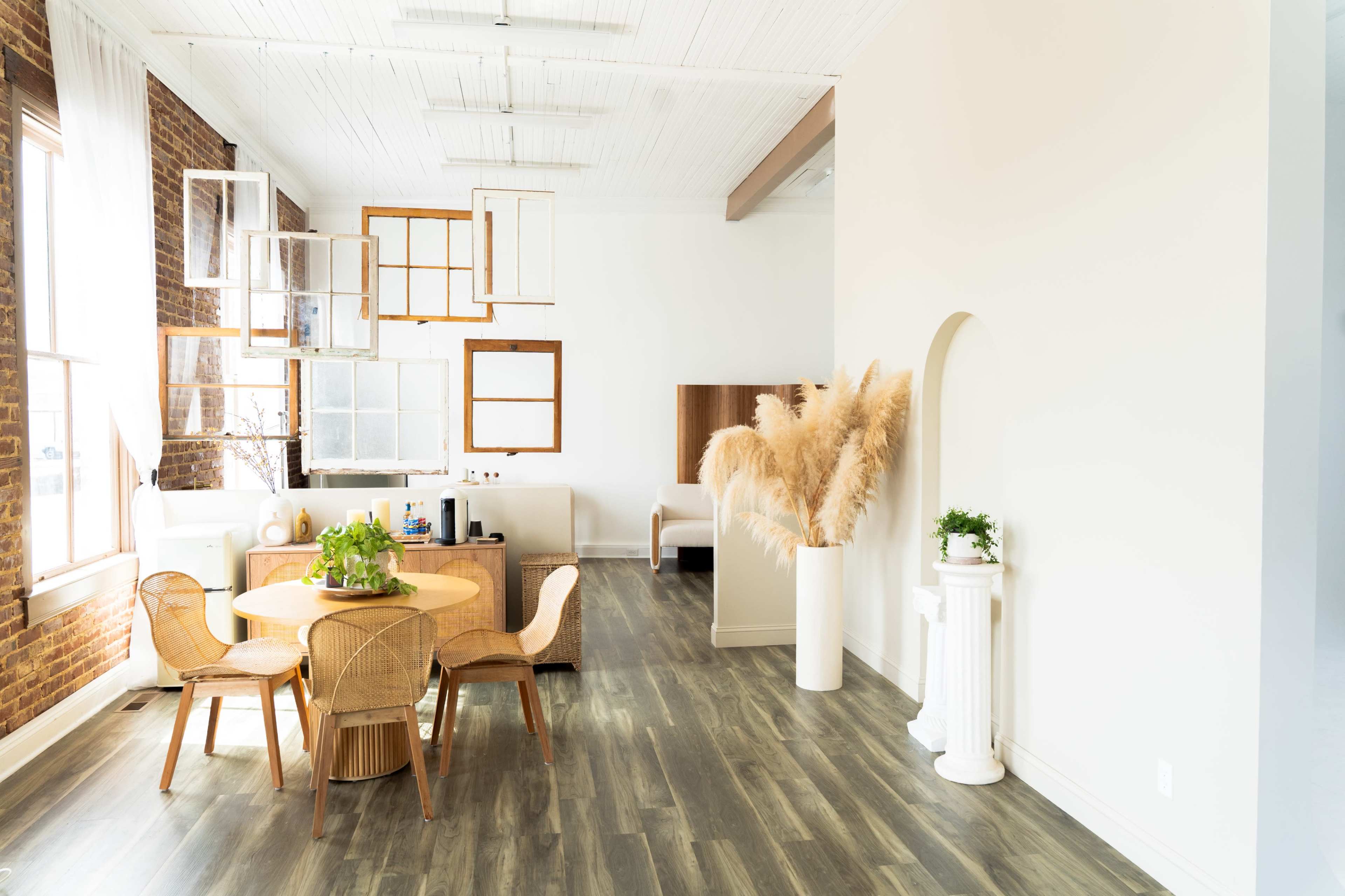 The image shows a bright, modern interior featuring a round dining table surrounded by wicker chairs, a large white plant arrangement, and various framed mirrors on the wall.
