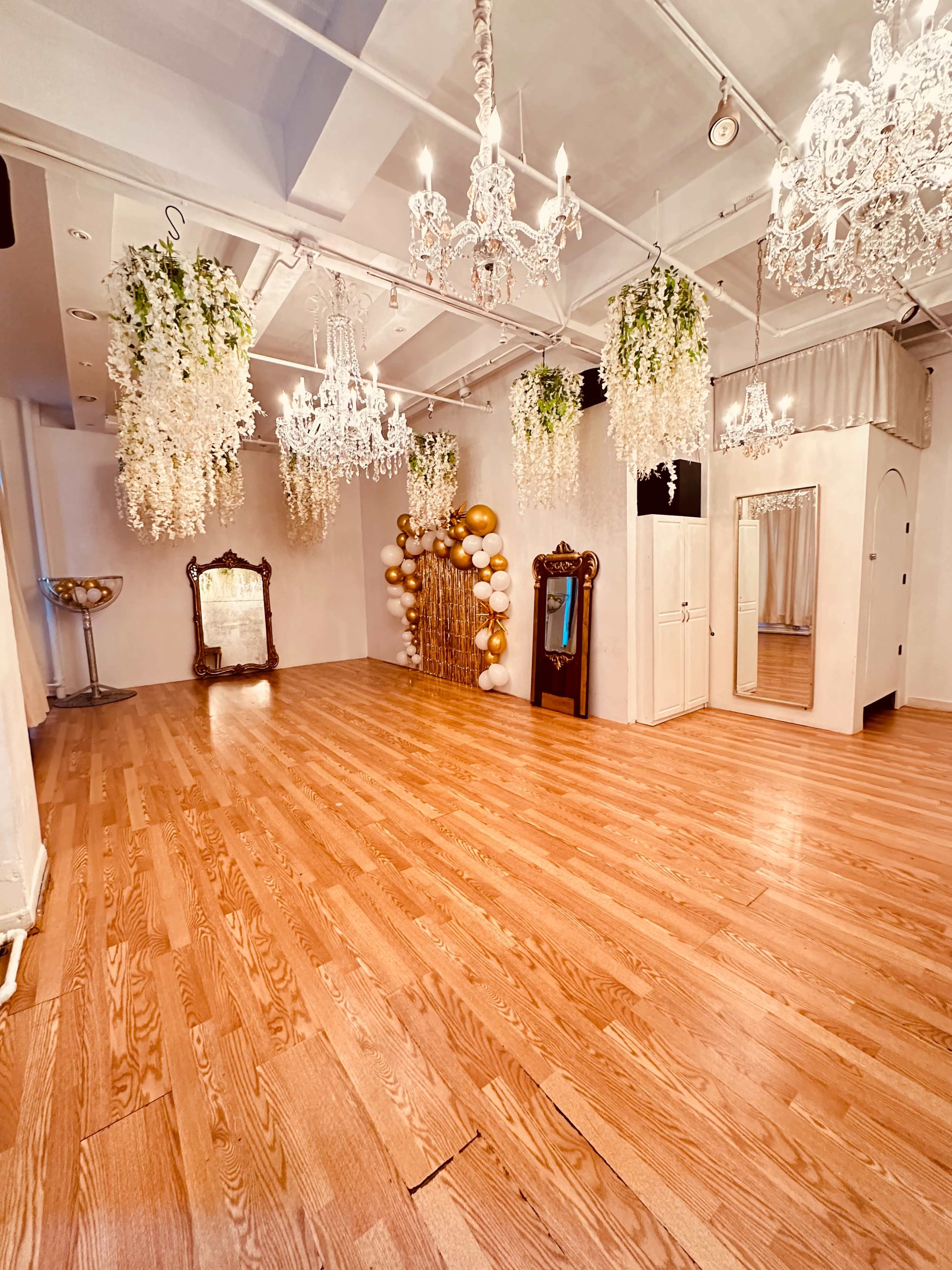 The image shows a spacious room adorned with chandeliers and decorated with floral arrangements and balloons, featuring a large mirror and wooden flooring.