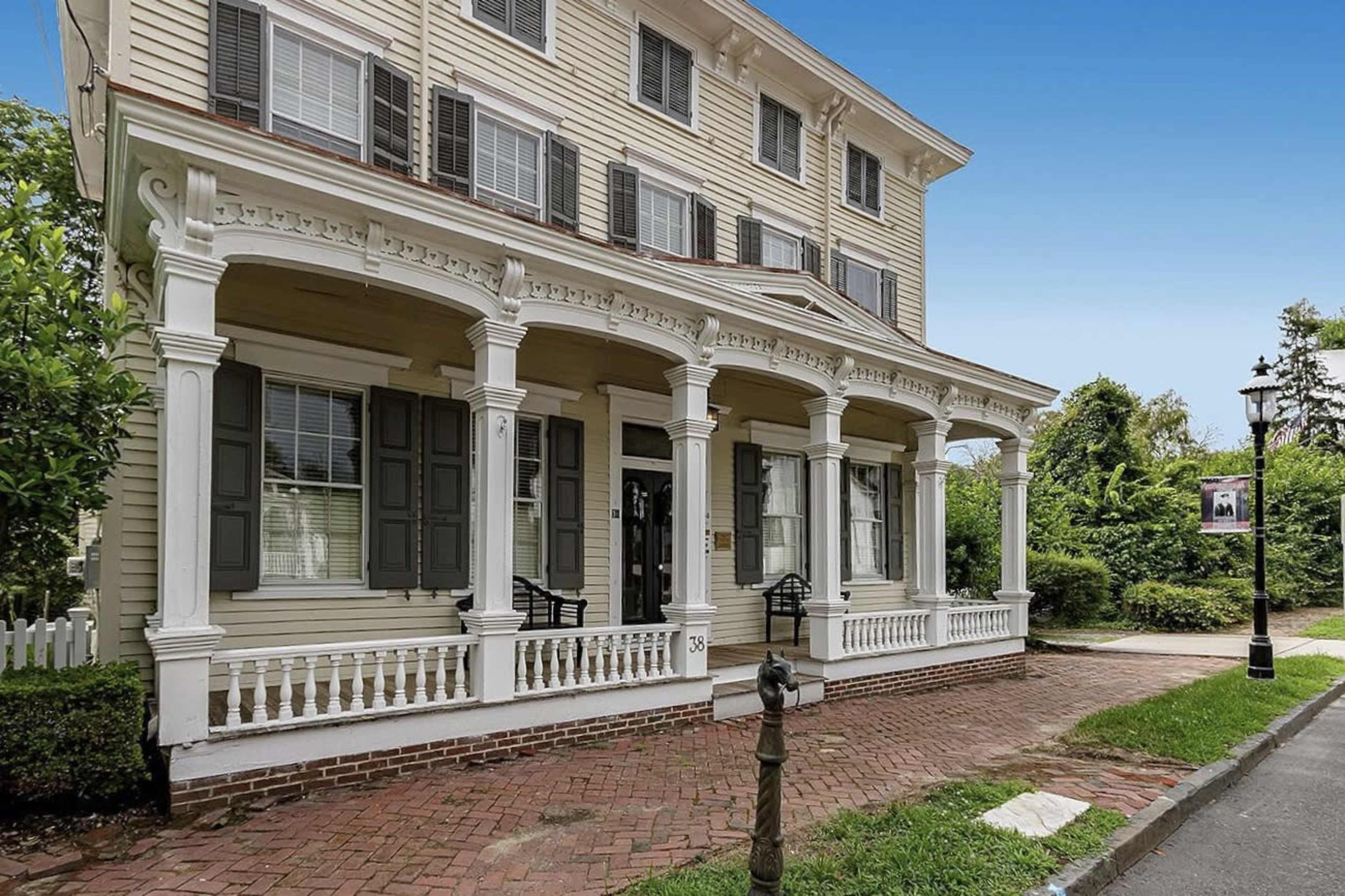 The image shows a three-story historical house with a large front porch supported by several white columns and green shutters on the windows.