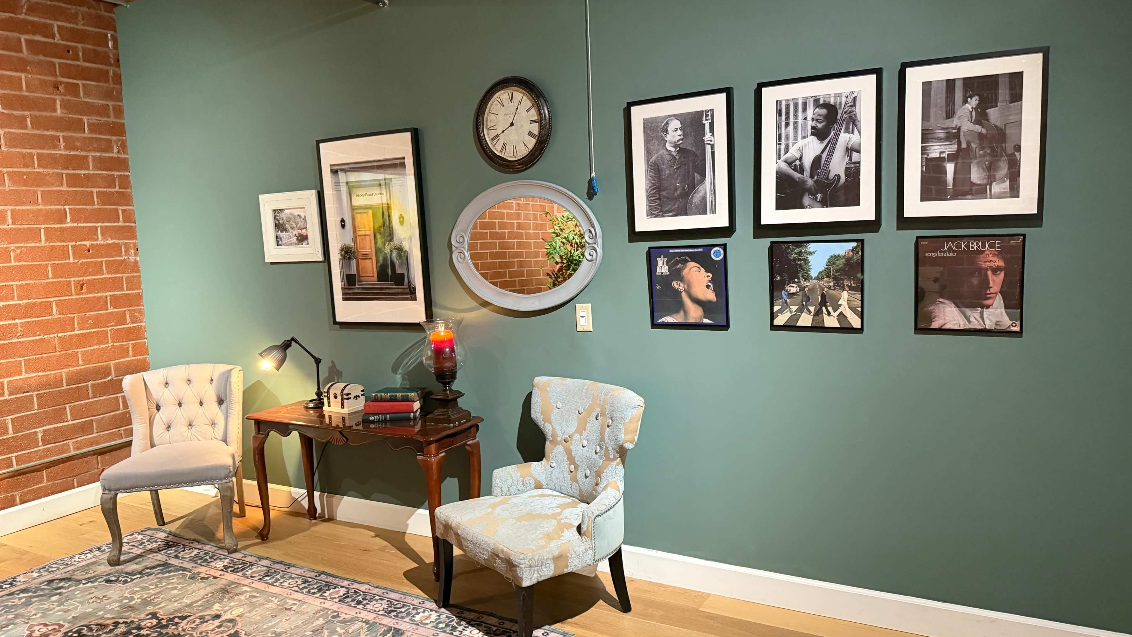 The image shows a cozy corner of a room featuring two chairs, a small table with a lamp, a wall clock, and a collection of framed black-and-white and color photographs hung on the green wall.