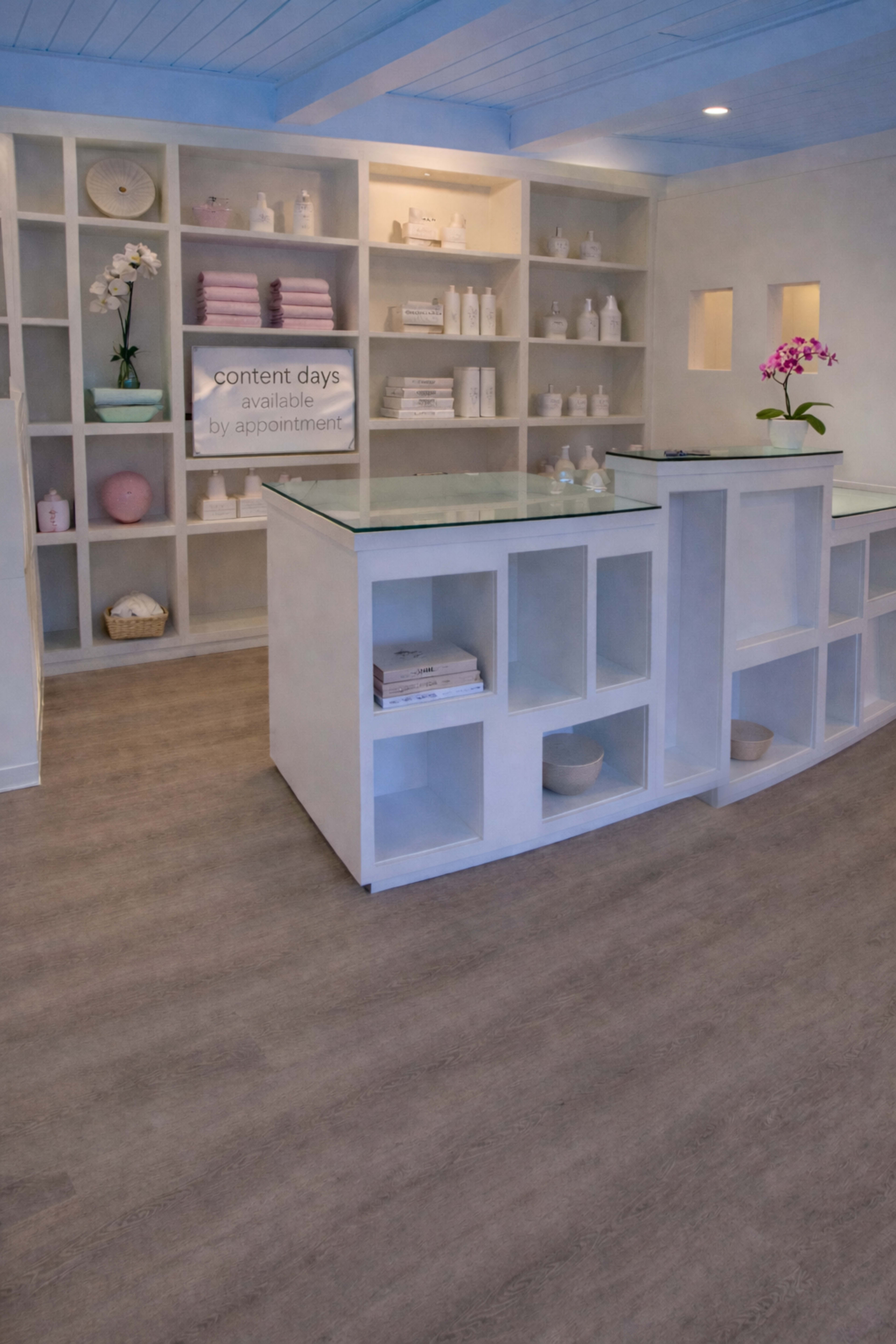 The image shows a modern reception area with a curved counter, organized shelving displaying various products, and light wood flooring.