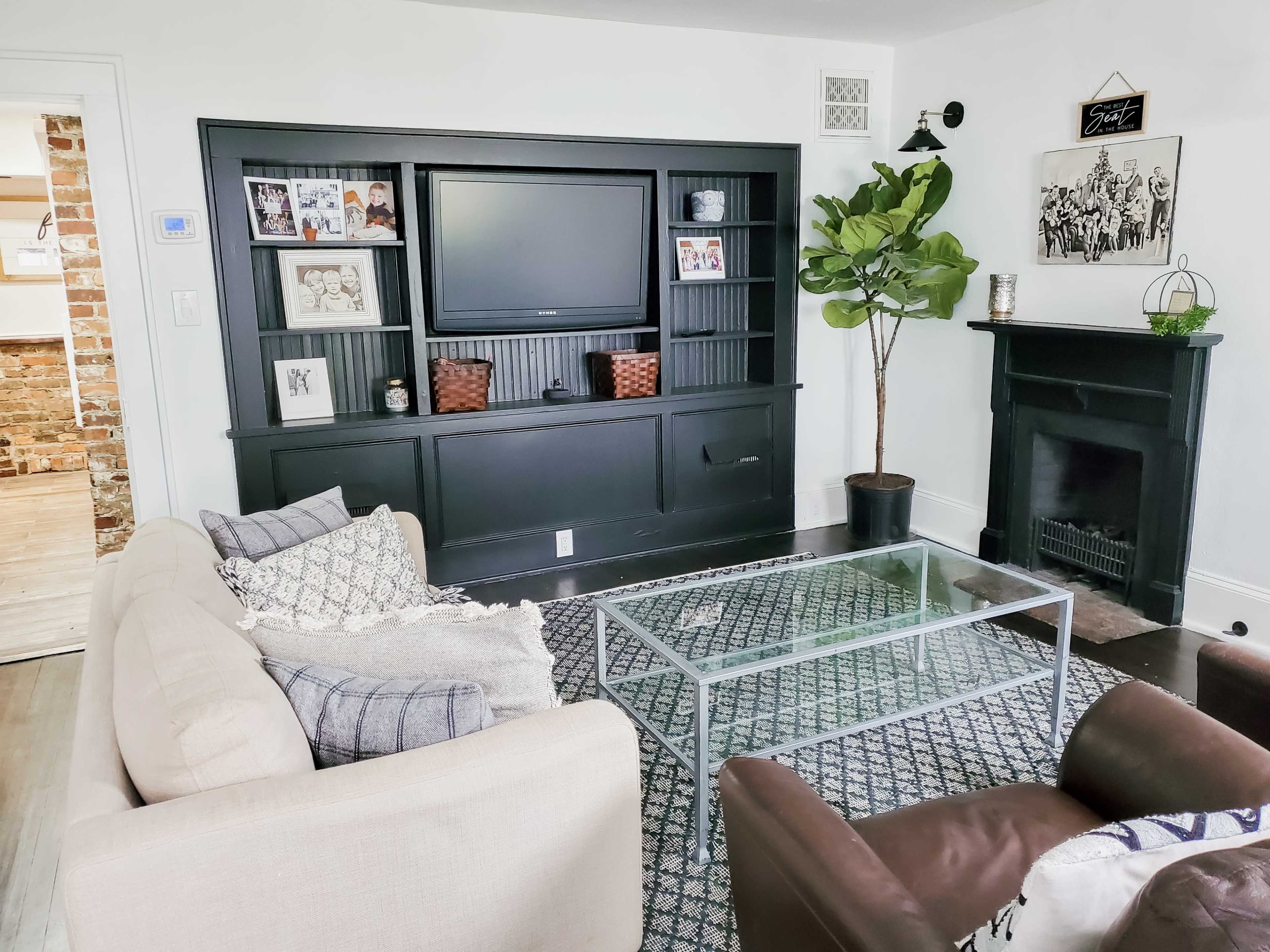 A living room with a black entertainment unit, a glass coffee table, a light-colored sofa, two brown armchairs, and a fireplace.