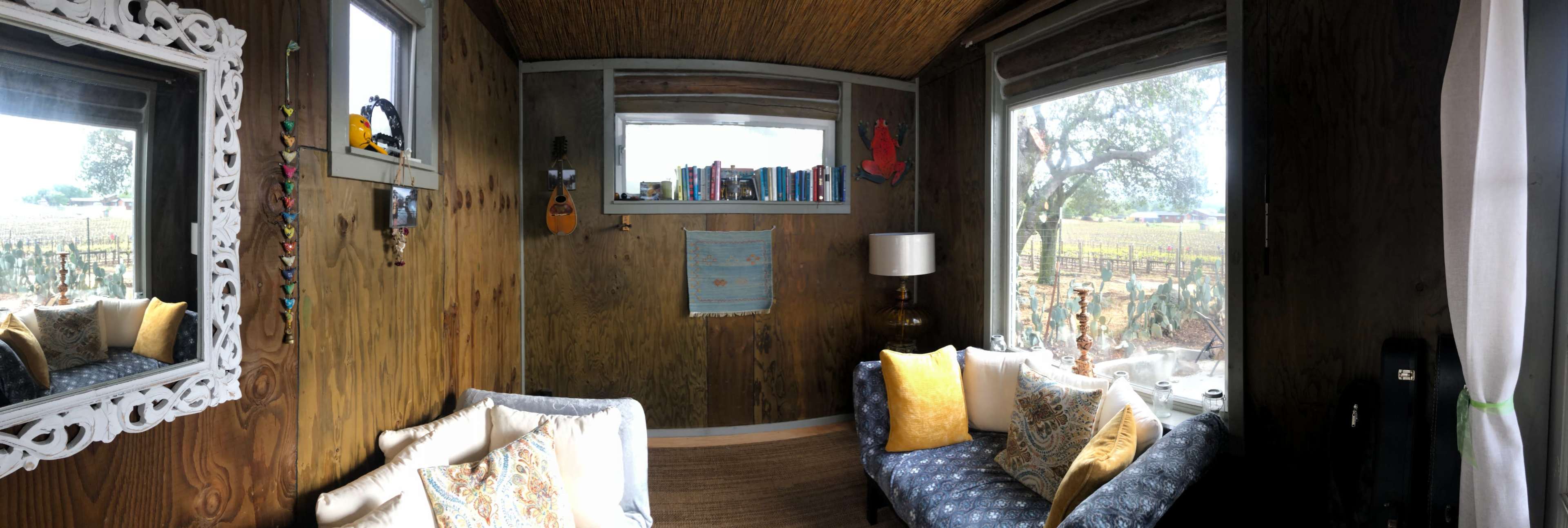The interior of a small, wooden room featuring a couch, a lamp, bookshelves, and large windows overlooking a vineyard.