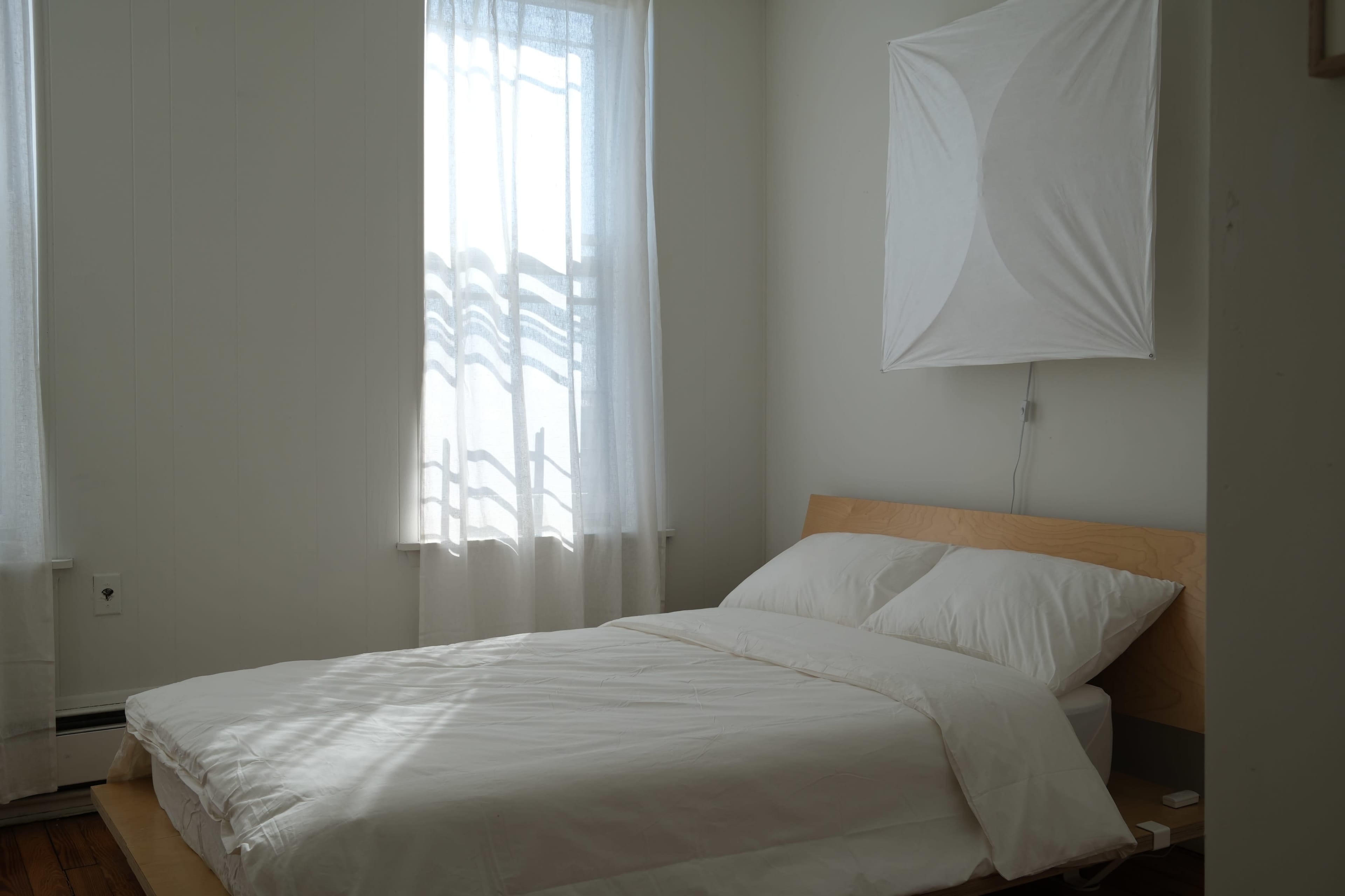 A minimalist bedroom features a bed with white linens, positioned beneath sheer curtains that allow natural light to filter in.