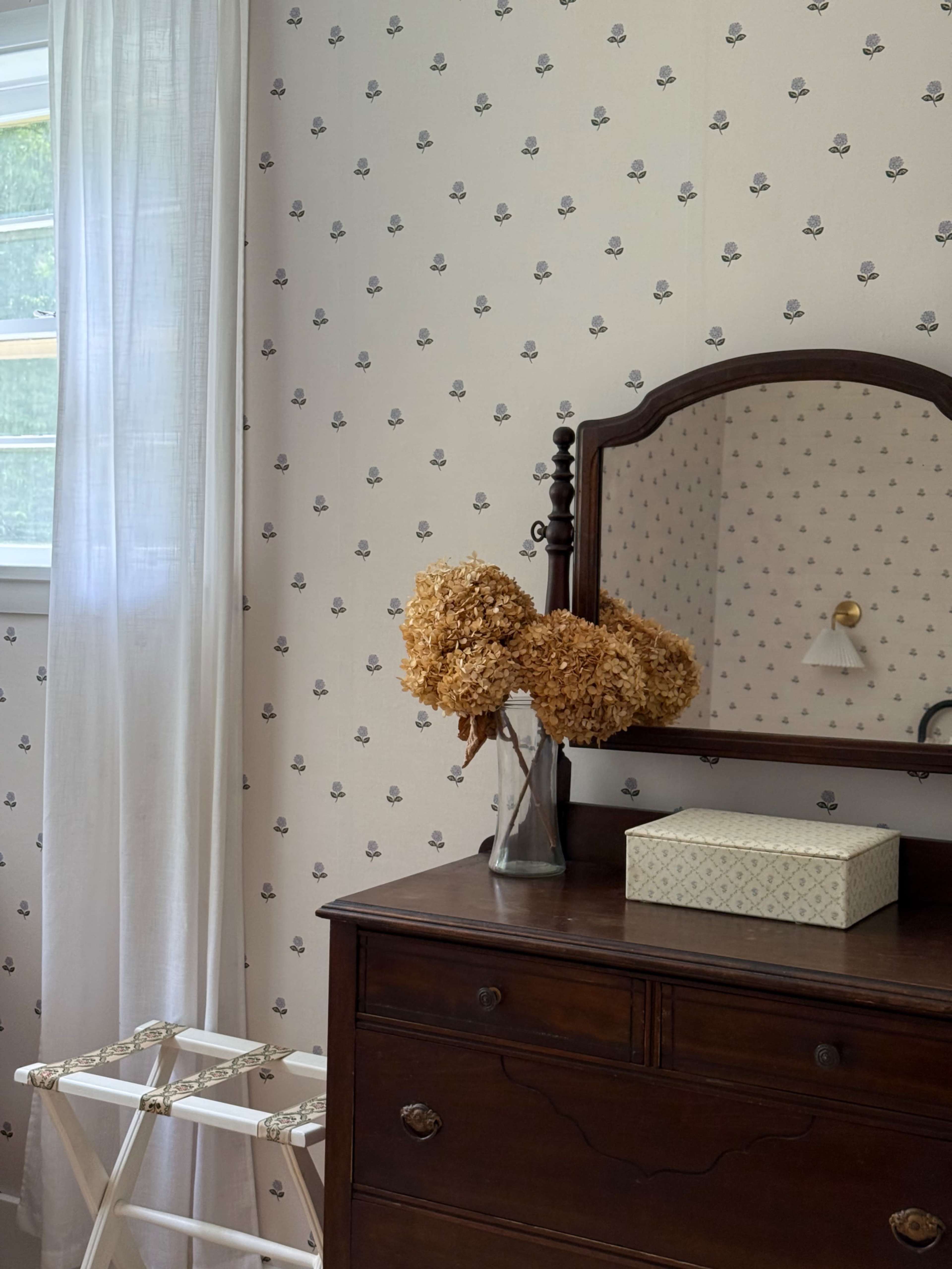 The image shows a corner of a bedroom featuring a dark wooden dresser, a vase of dried flowers, a decorative box, a mirror, and patterned wallpaper.