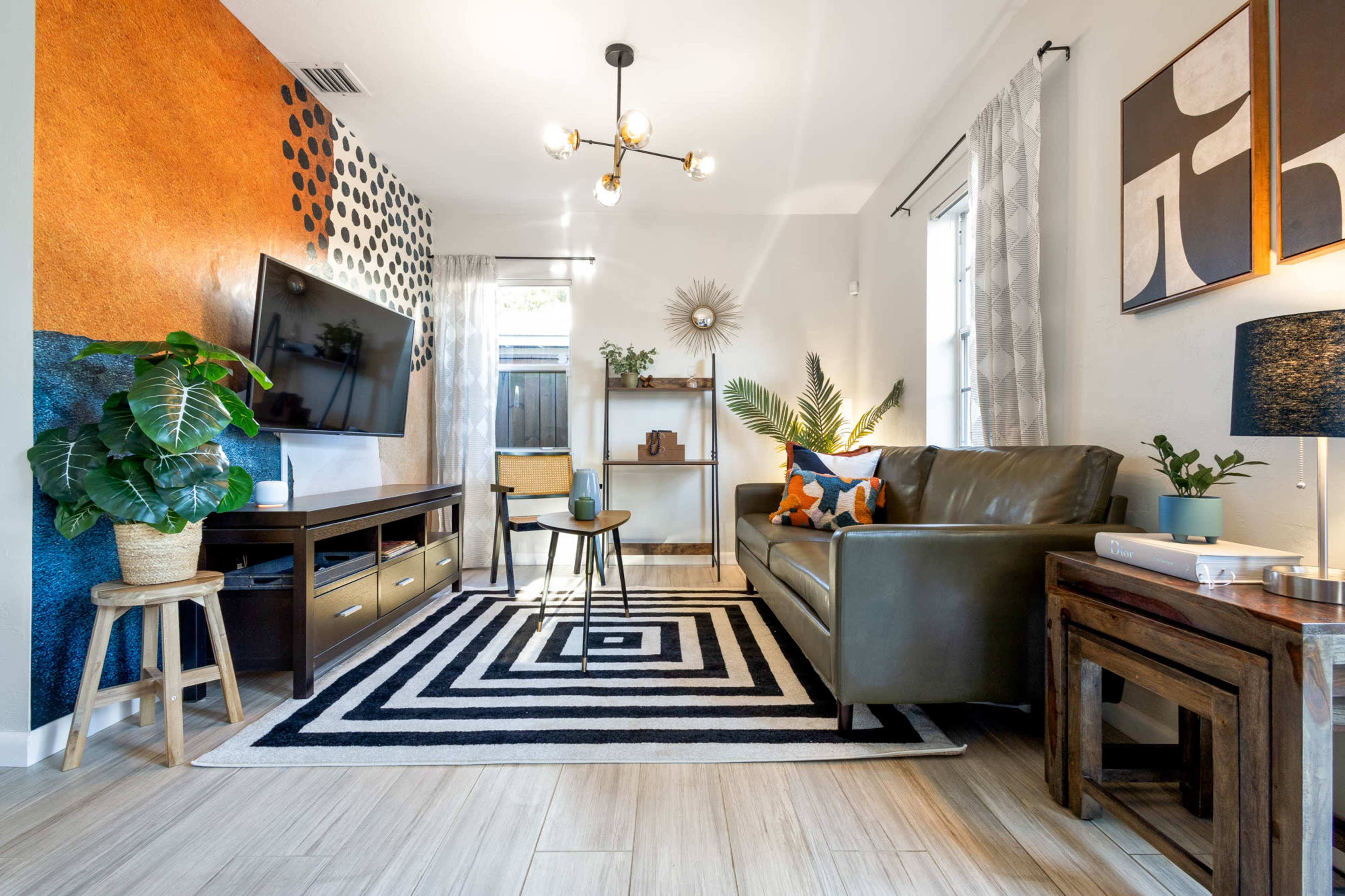 A modern living room features a dark sofa, a geometric area rug, a large television, and decorative plants against a backdrop of textured wall paint.