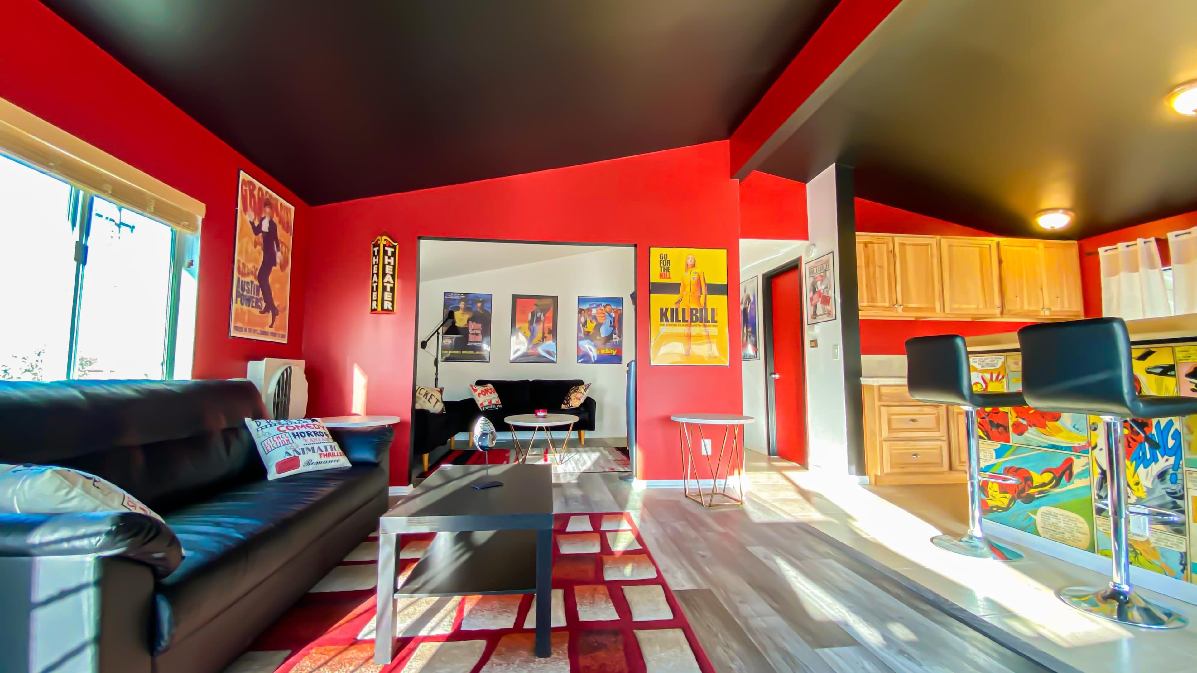 The image shows a living room with red walls, a black leather sofa, a glass coffee table, and vibrant movie posters on the walls.