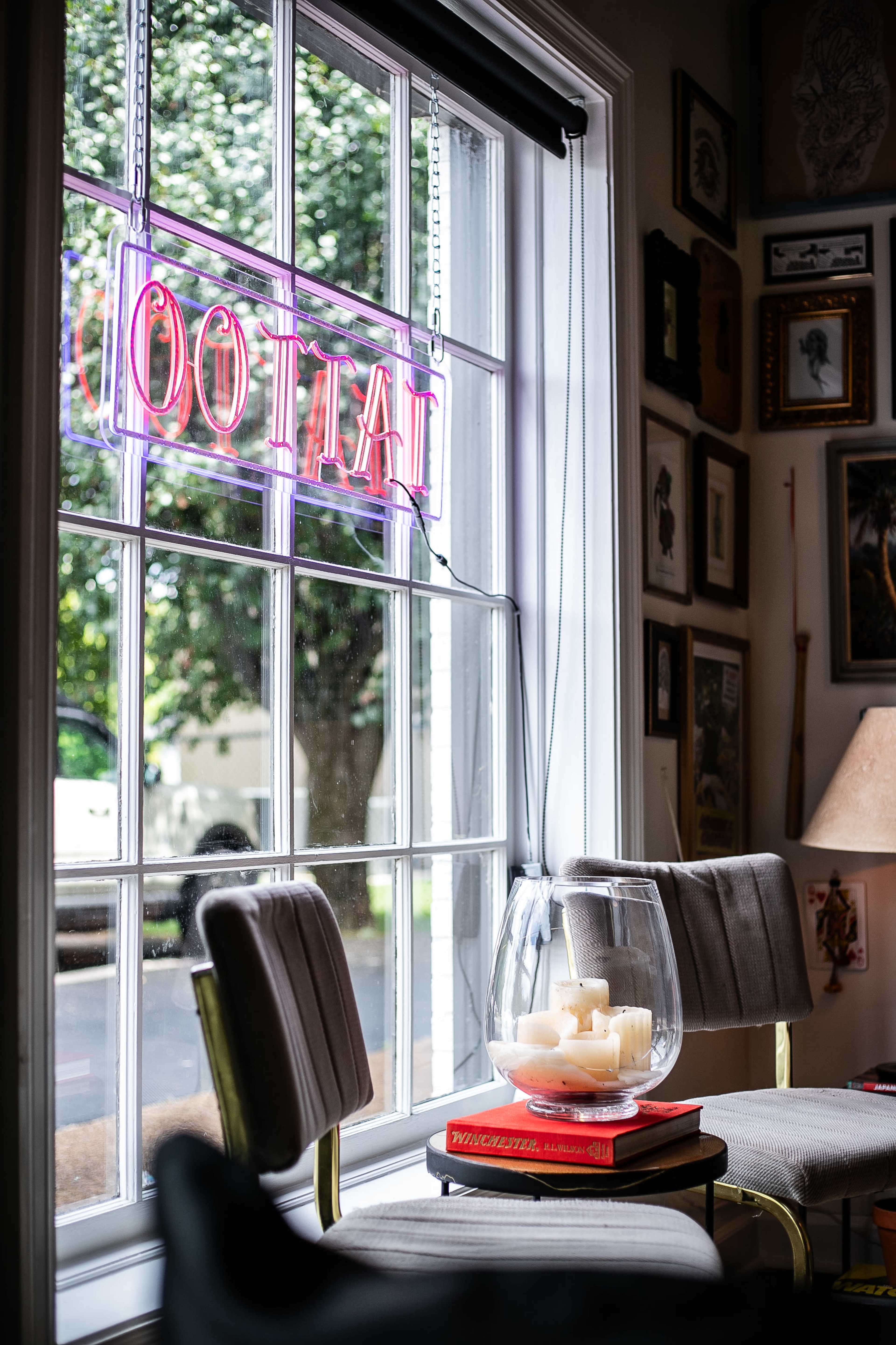 A window displays a neon "TATTOO" sign, alongside two chairs and a glass bowl of candles on a stack of books.