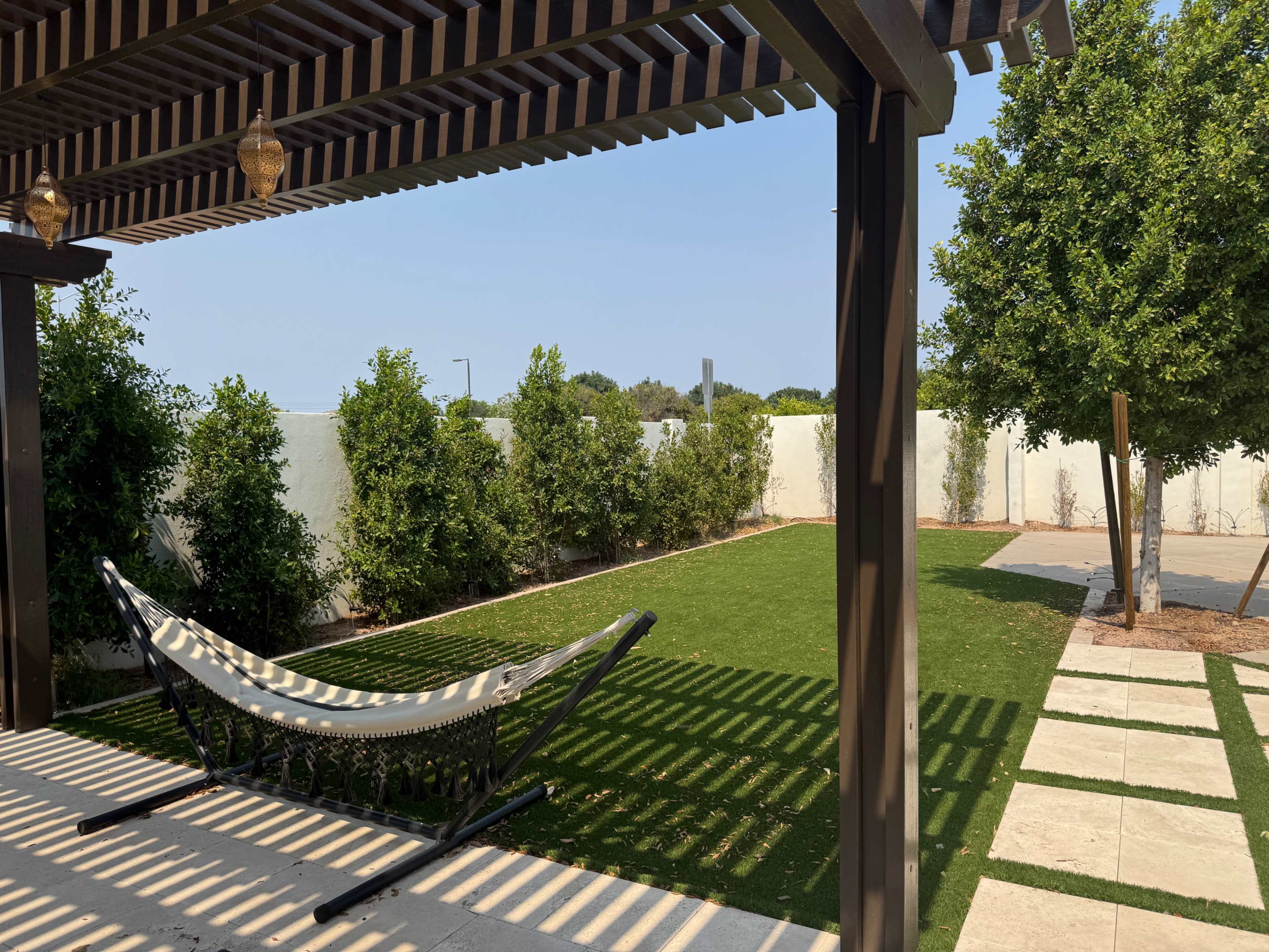 A hammock is suspended under a wooden pergola in a landscaped yard with grass and shrubs.