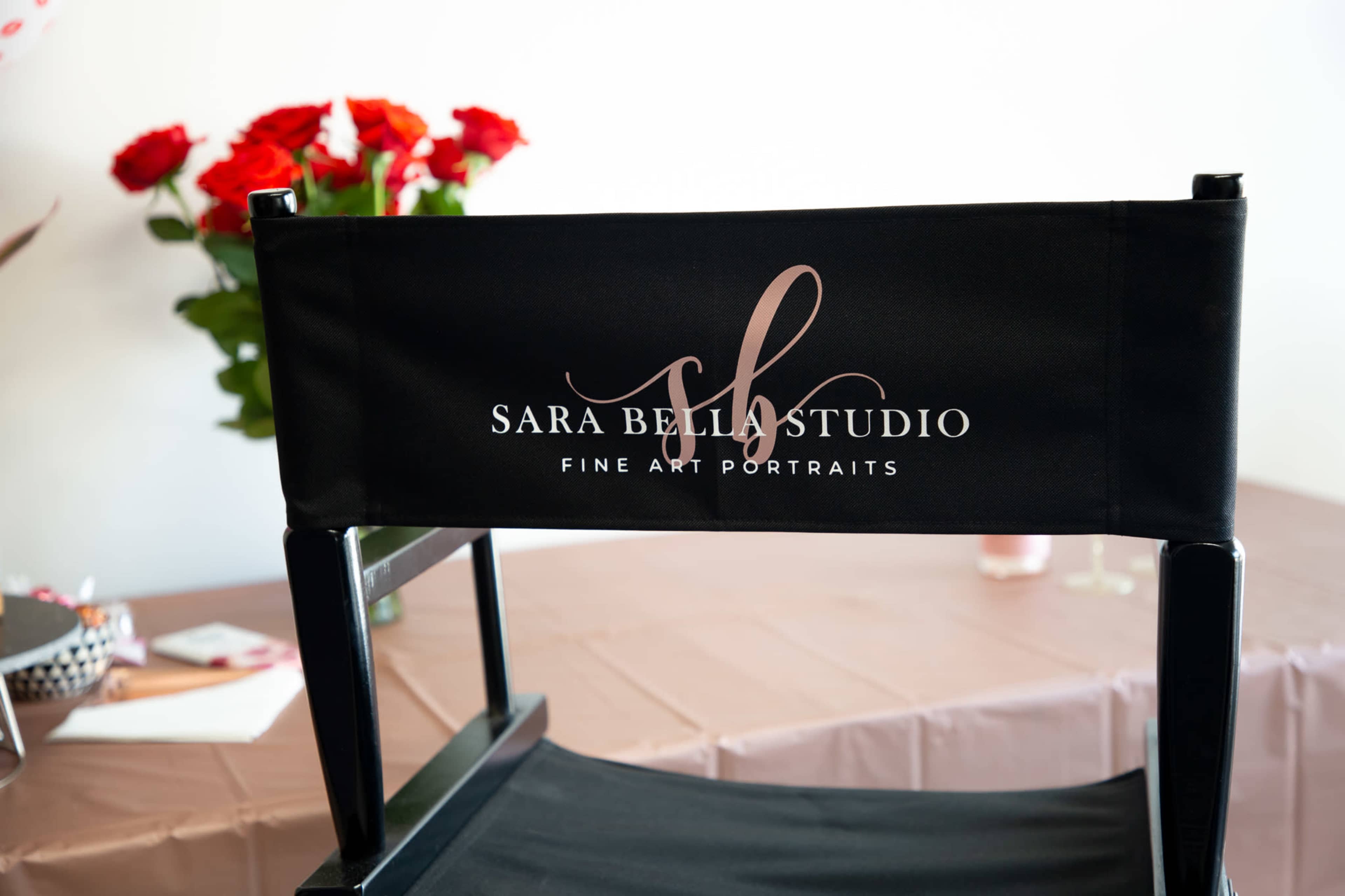 A black director's chair with the logo of "Sara Bella Studio" is positioned in front of a table decorated with a brown tablecloth and vases of red roses.