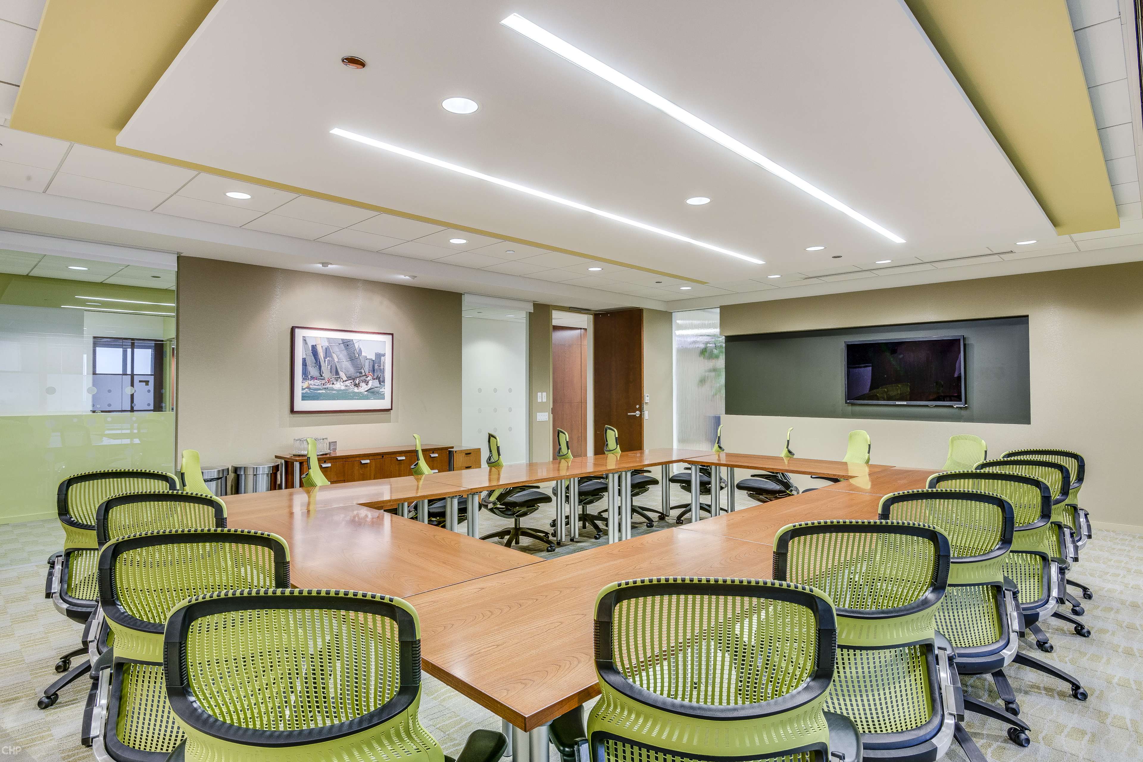 A modern conference room features a large table surrounded by green ergonomic chairs, with a television mounted on the wall and natural light coming from windows.