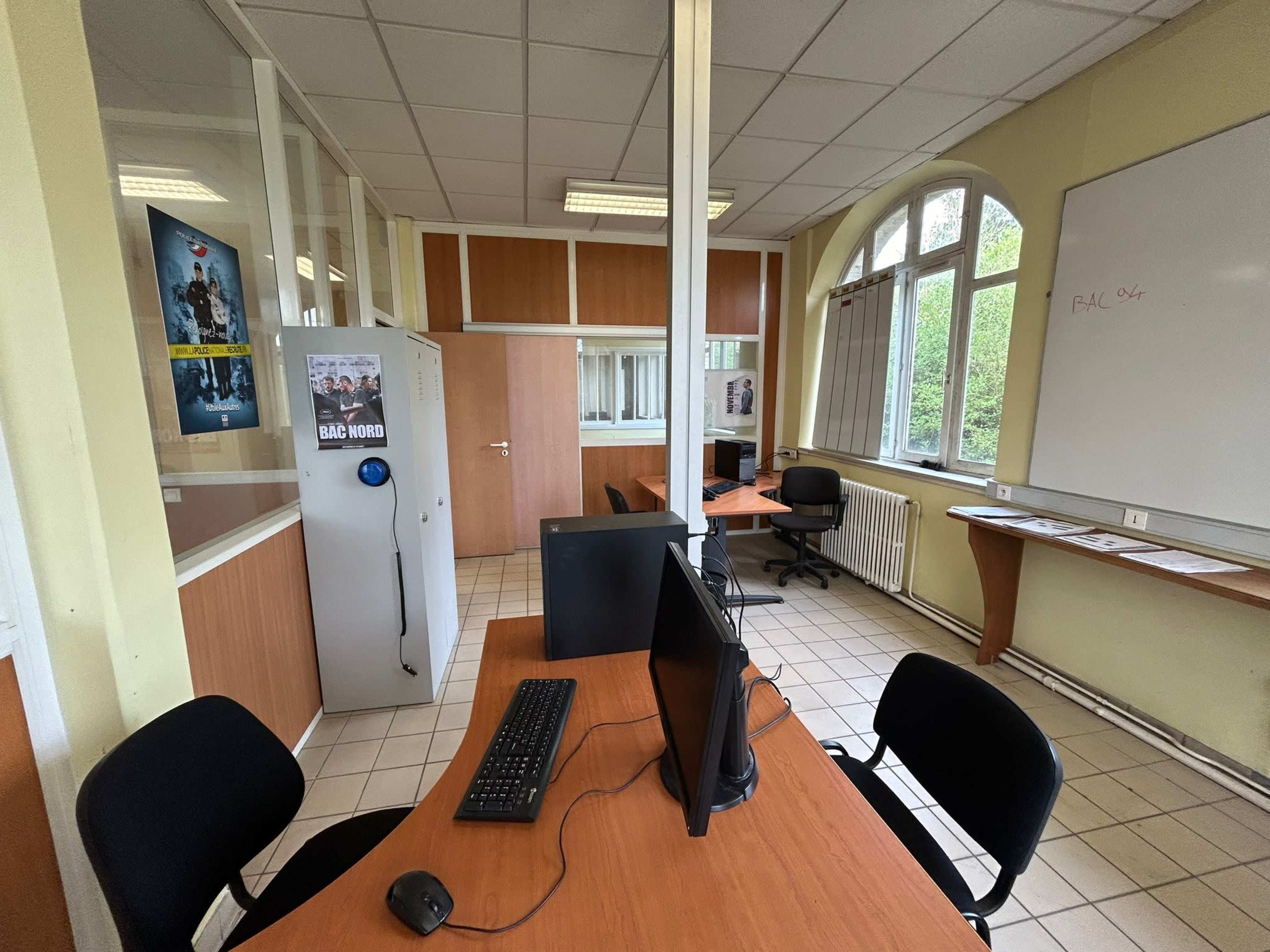 An office space with a computer setup, desks, a refrigerator, and large windows allowing natural light.