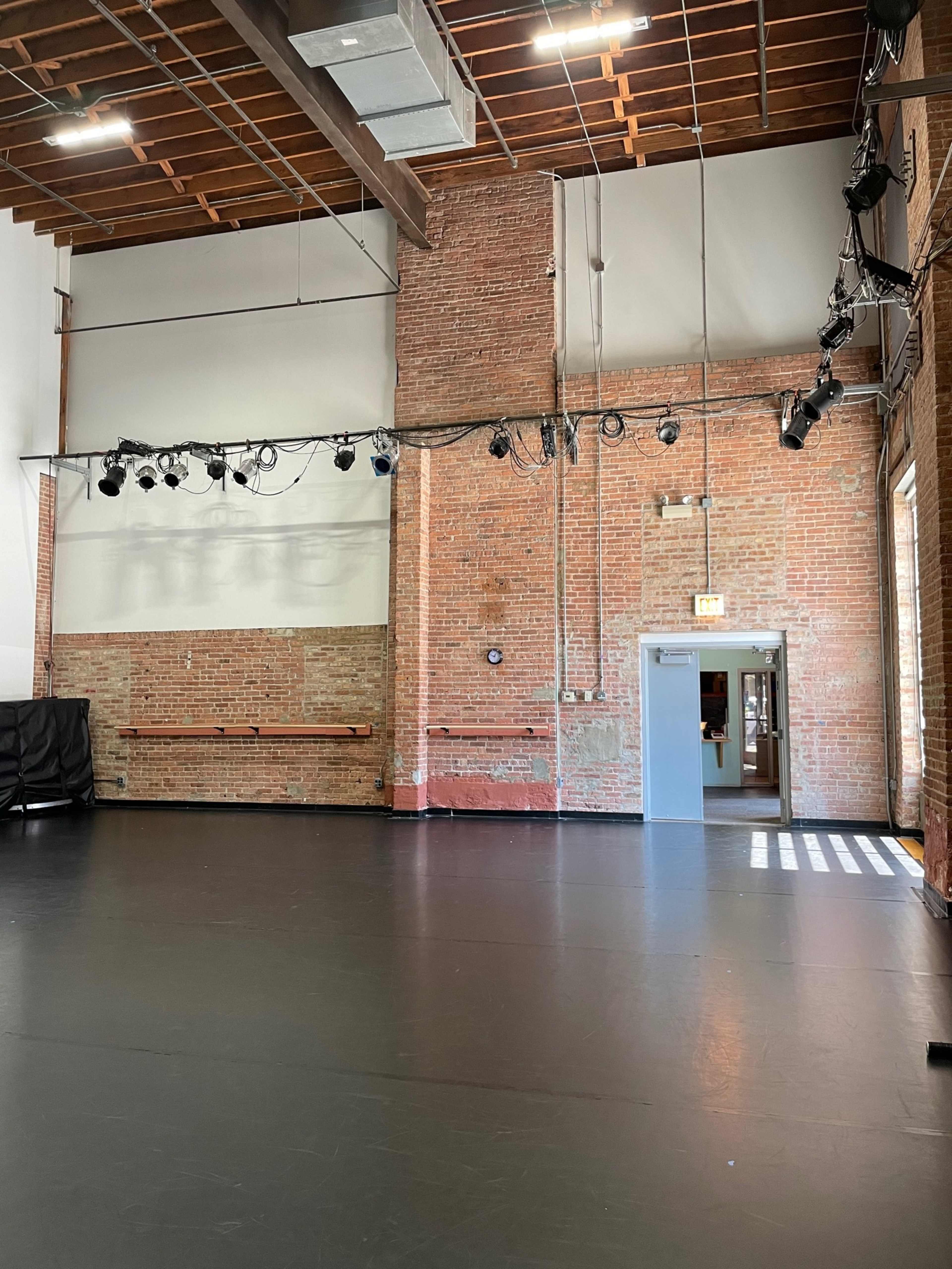 The image shows a spacious rehearsal studio with exposed brick walls, a polished black floor, and a row of overhead lights.