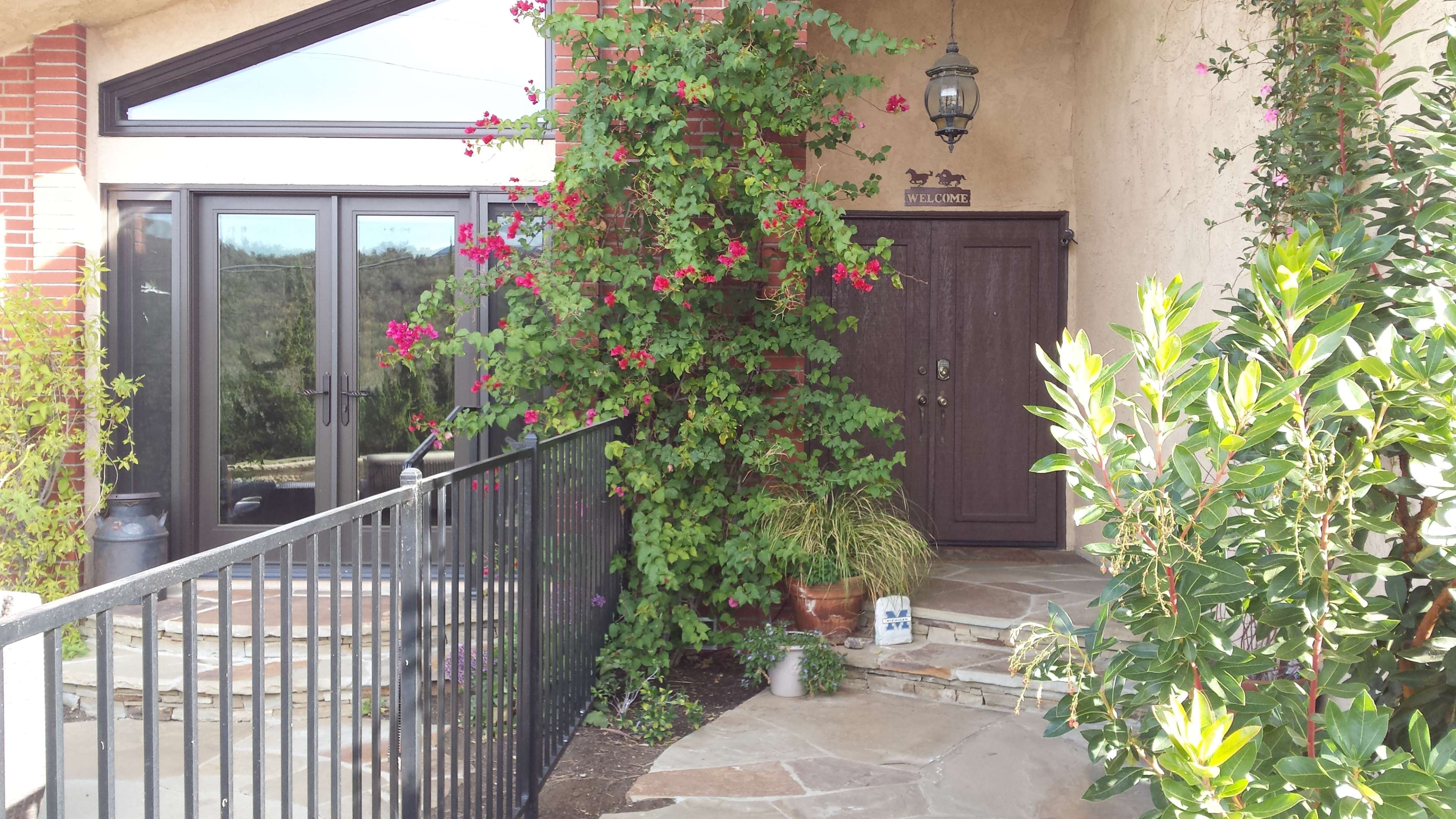A pathway leads to a front door surrounded by flowering plants and a small welcome sign.