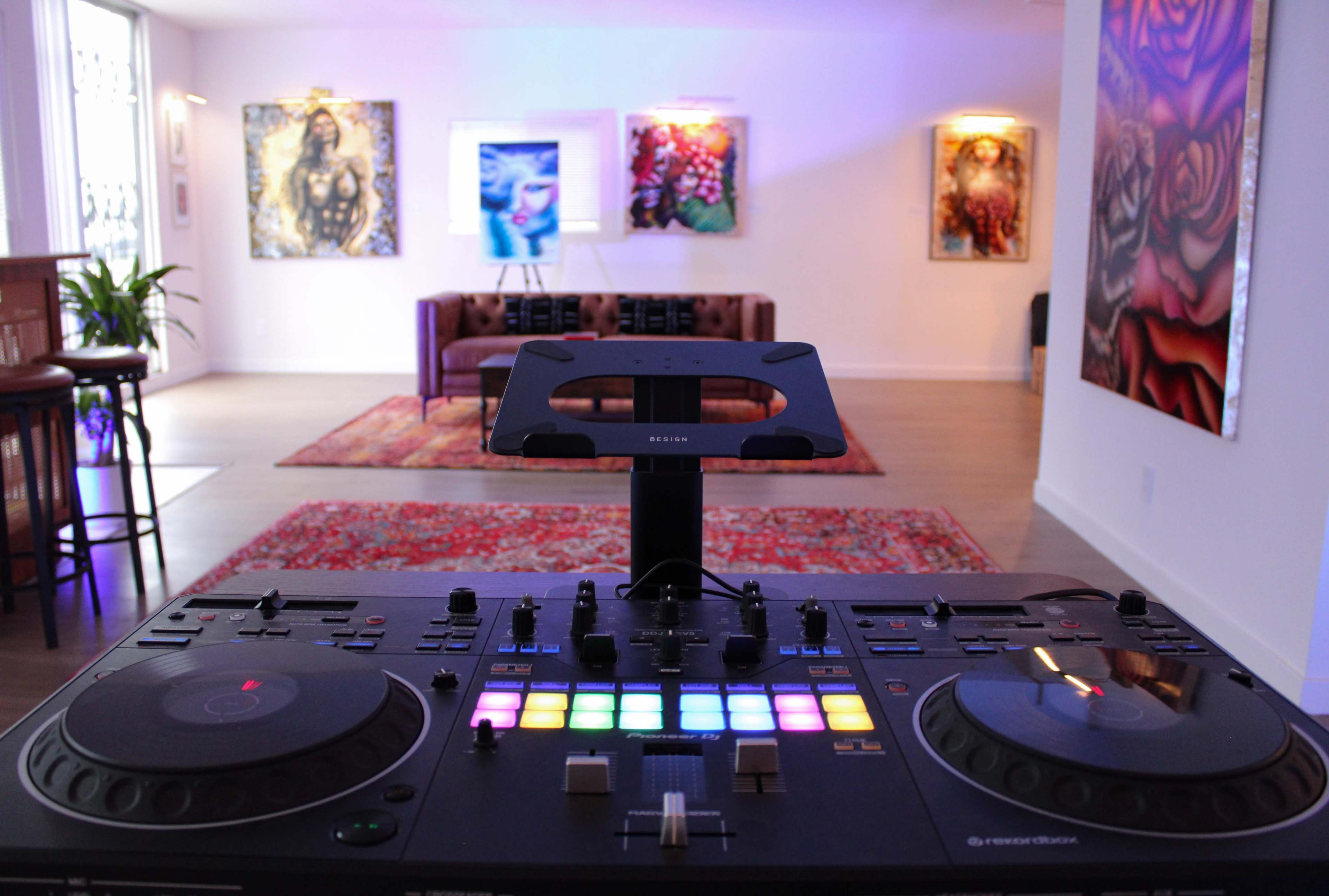 The image shows a DJ console in the foreground with colorful lights, facing an artistic room featuring a couch and several vibrant paintings on the walls.