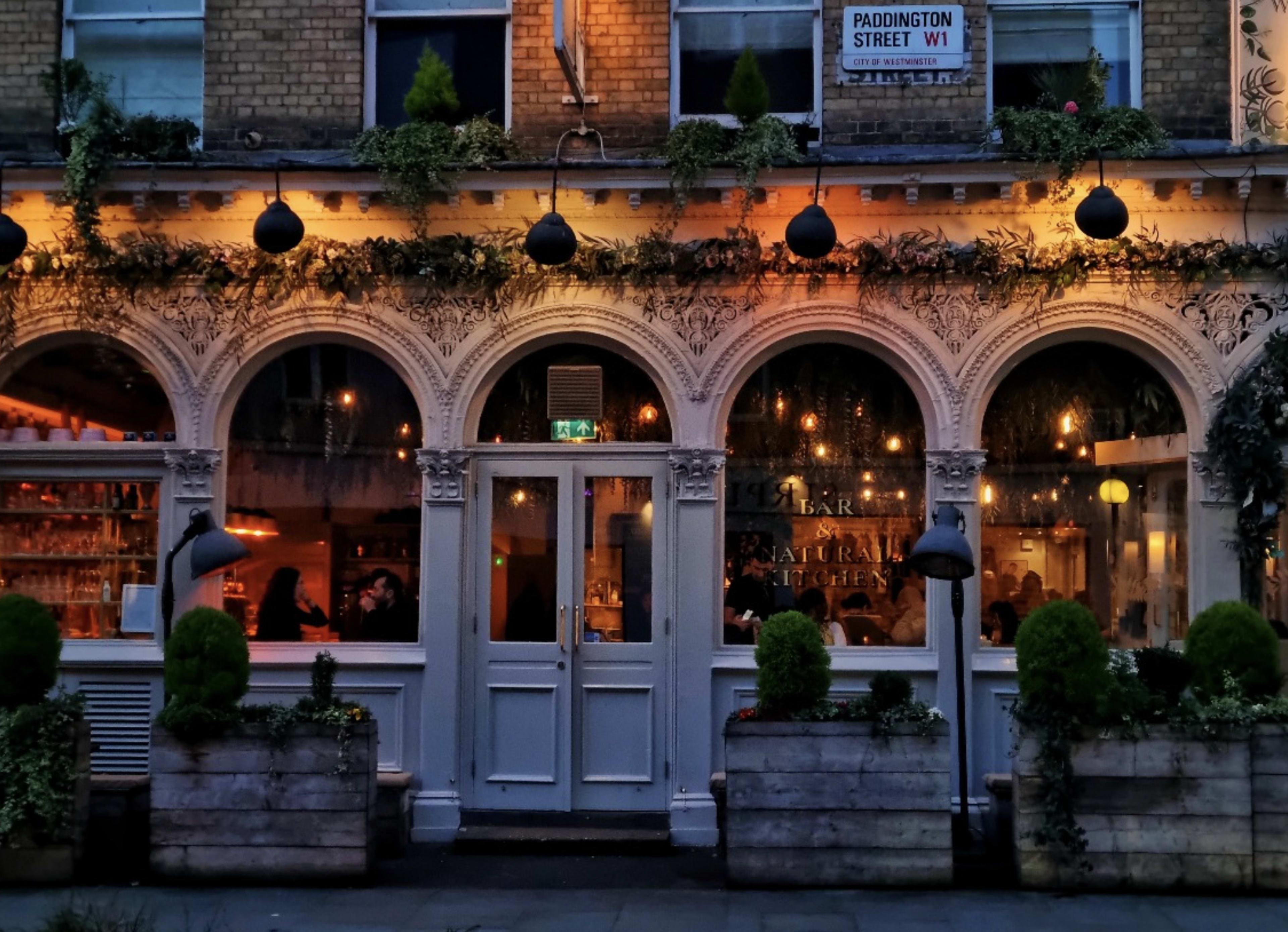 The image depicts a restaurant with large arched windows, adorned with outdoor plants, and illuminated by warm lights on Paddington Street.