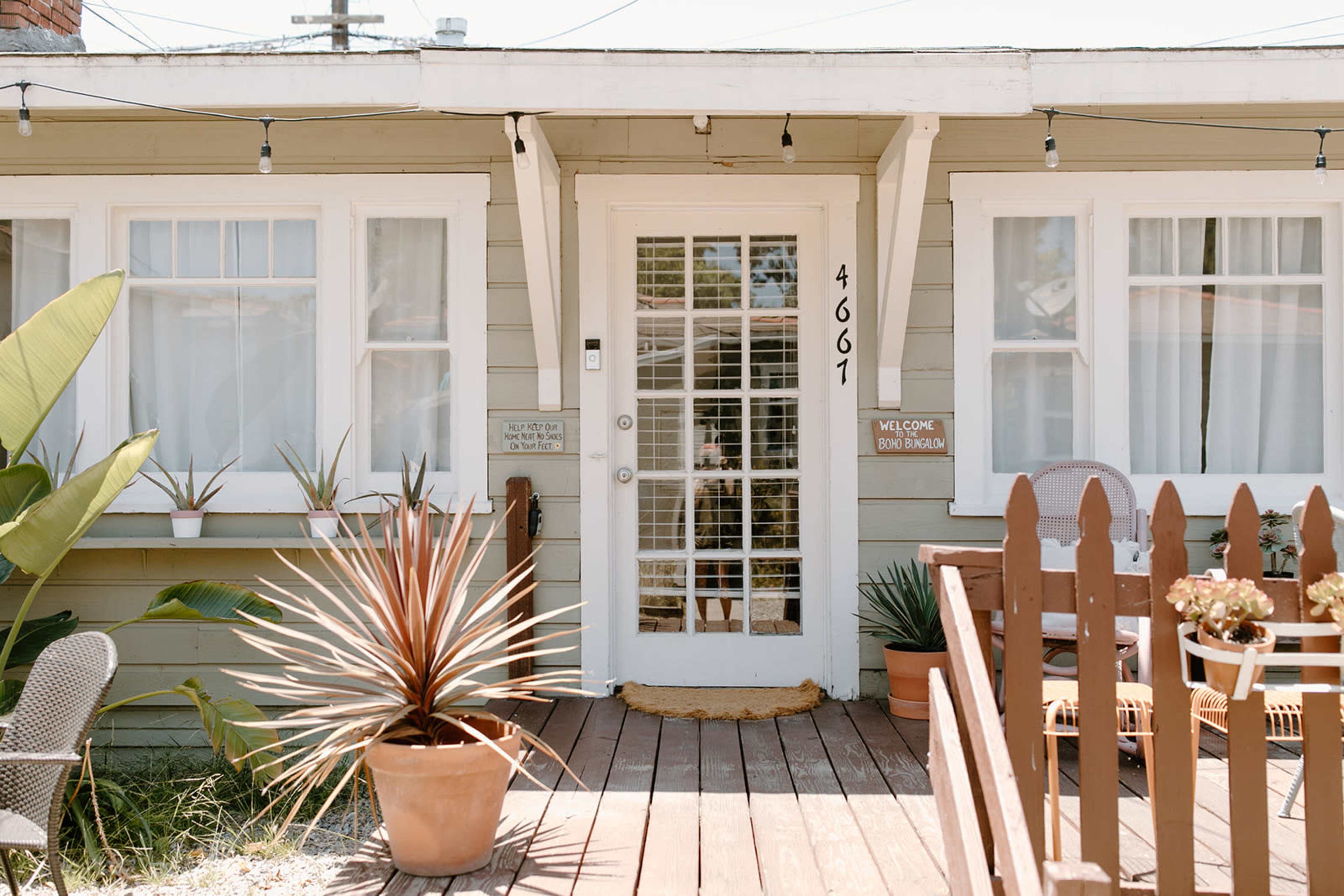 A light-colored house with a front porch, featuring a white door, potted plants, and wooden fencing.
