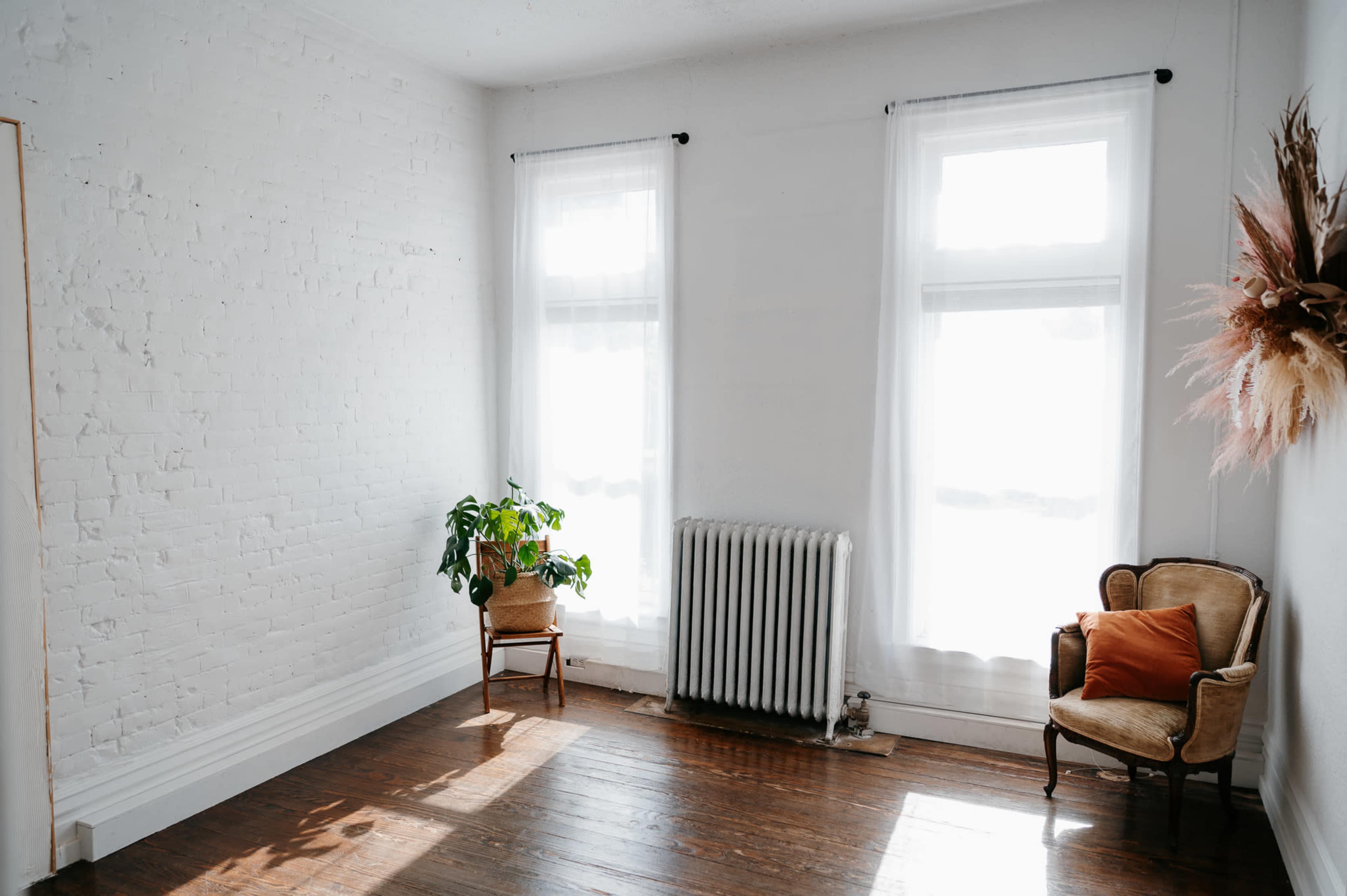 A sunlit room with white brick walls, two large windows, a potted plant, a small chair, and a radiator.