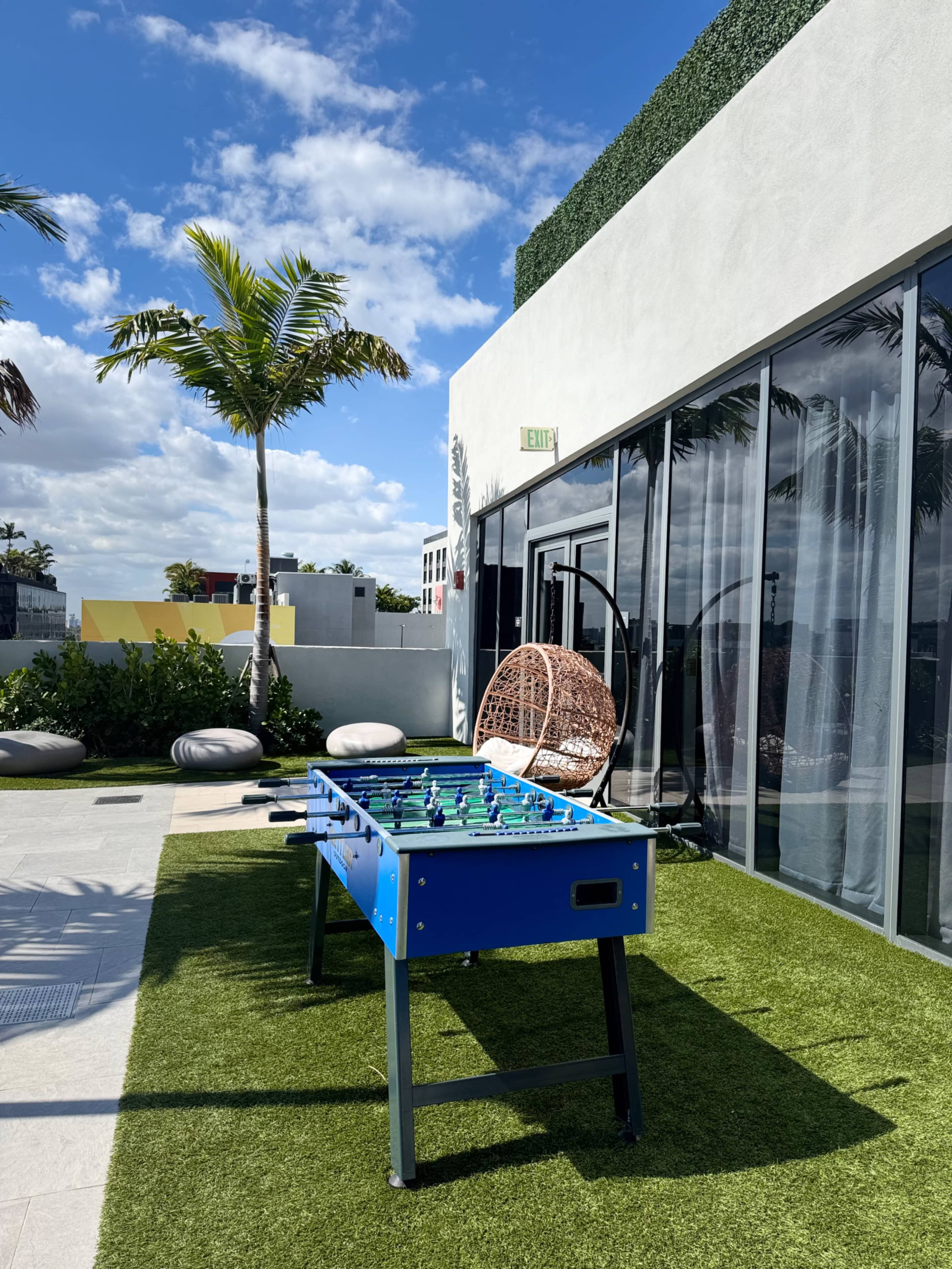 A blue foosball table is set on green artificial grass near a modern building with large glass windows and palm trees in the background.