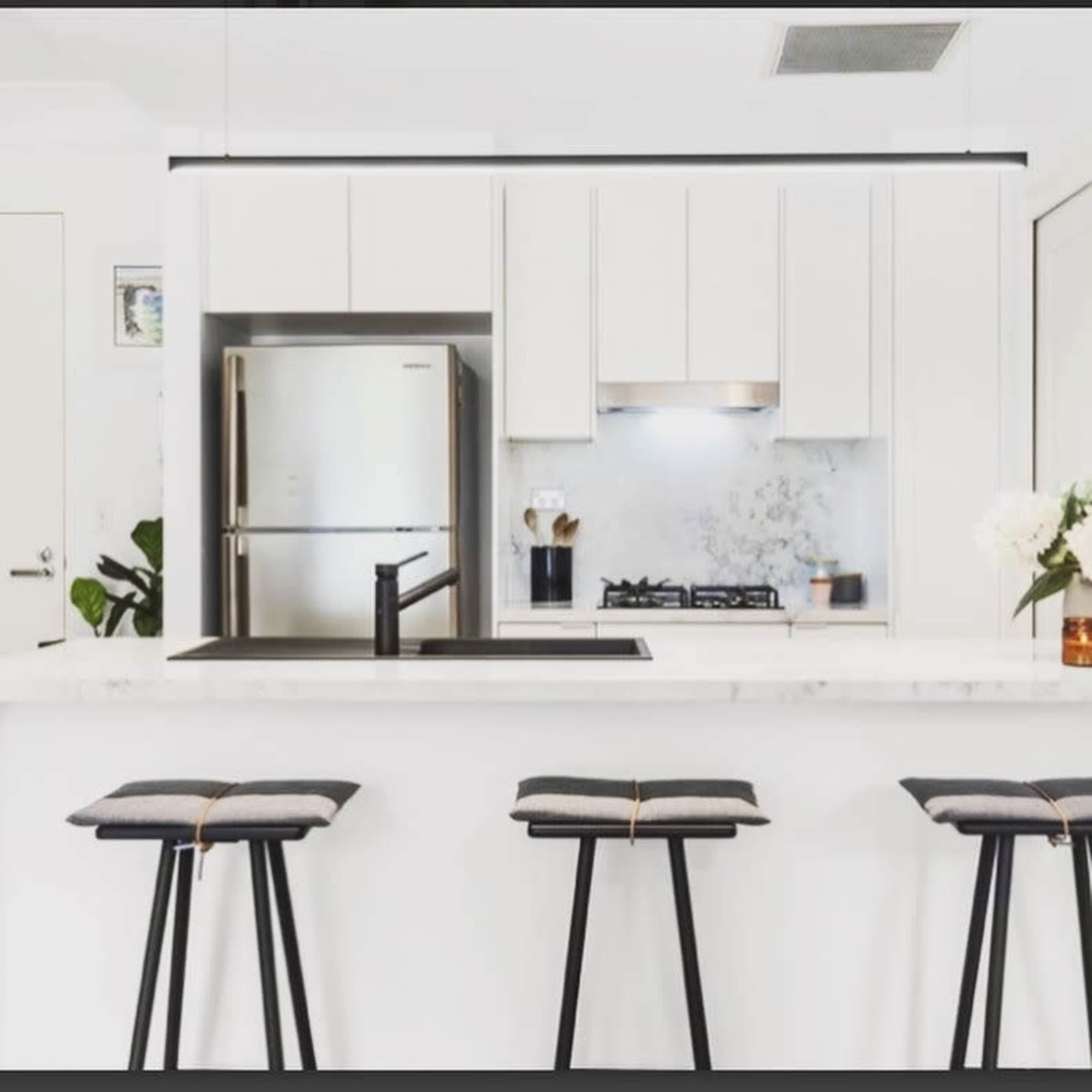 A modern kitchen features a countertop with three black stools, a stainless steel refrigerator, and white cabinetry with a marble backsplash.