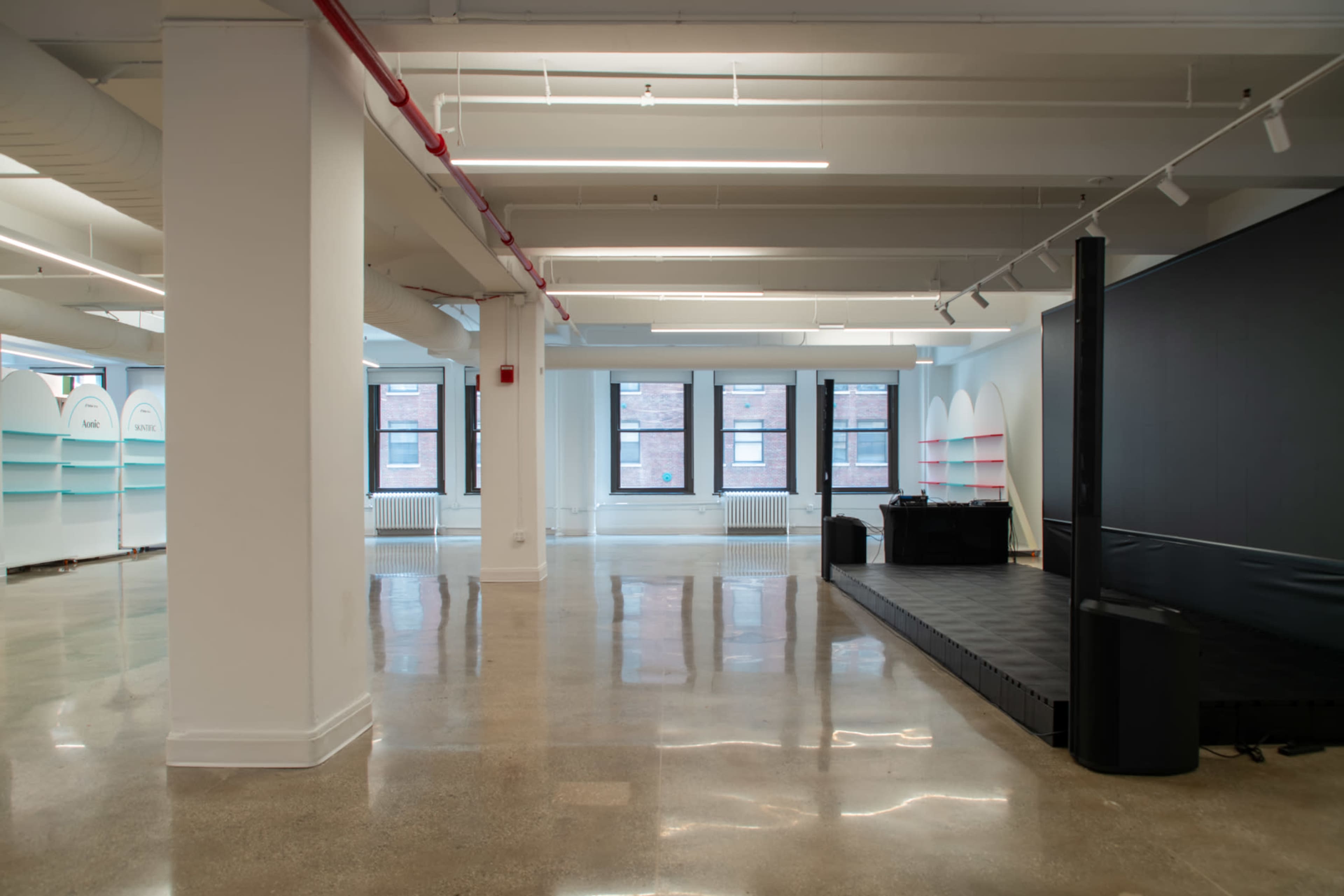 The image shows a spacious, modern interior with polished concrete flooring, large windows allowing natural light, and a small stage area in one corner.
