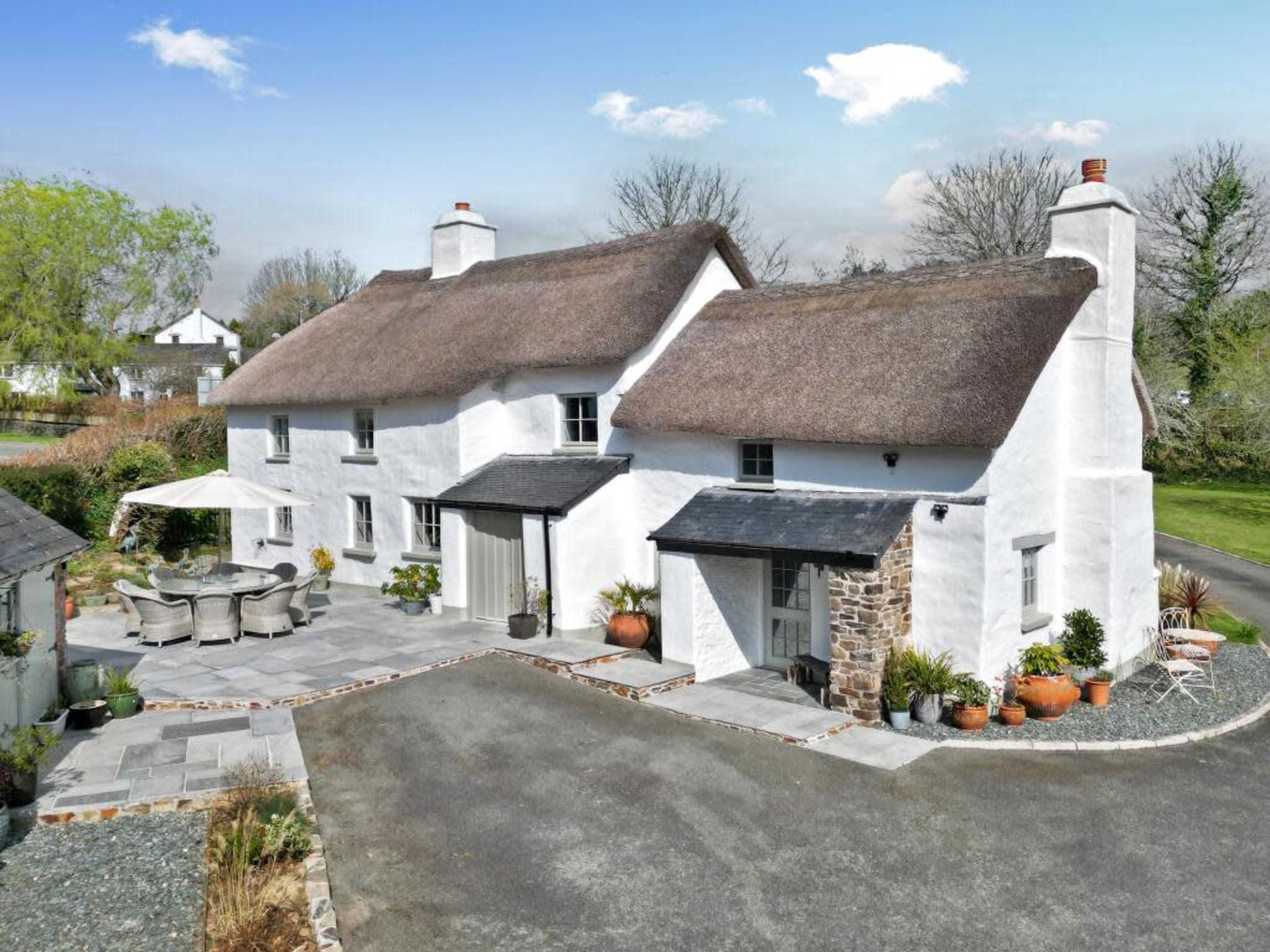The image shows a white, thatched-roof cottage surrounded by a stone patio, garden, and outdoor seating area.