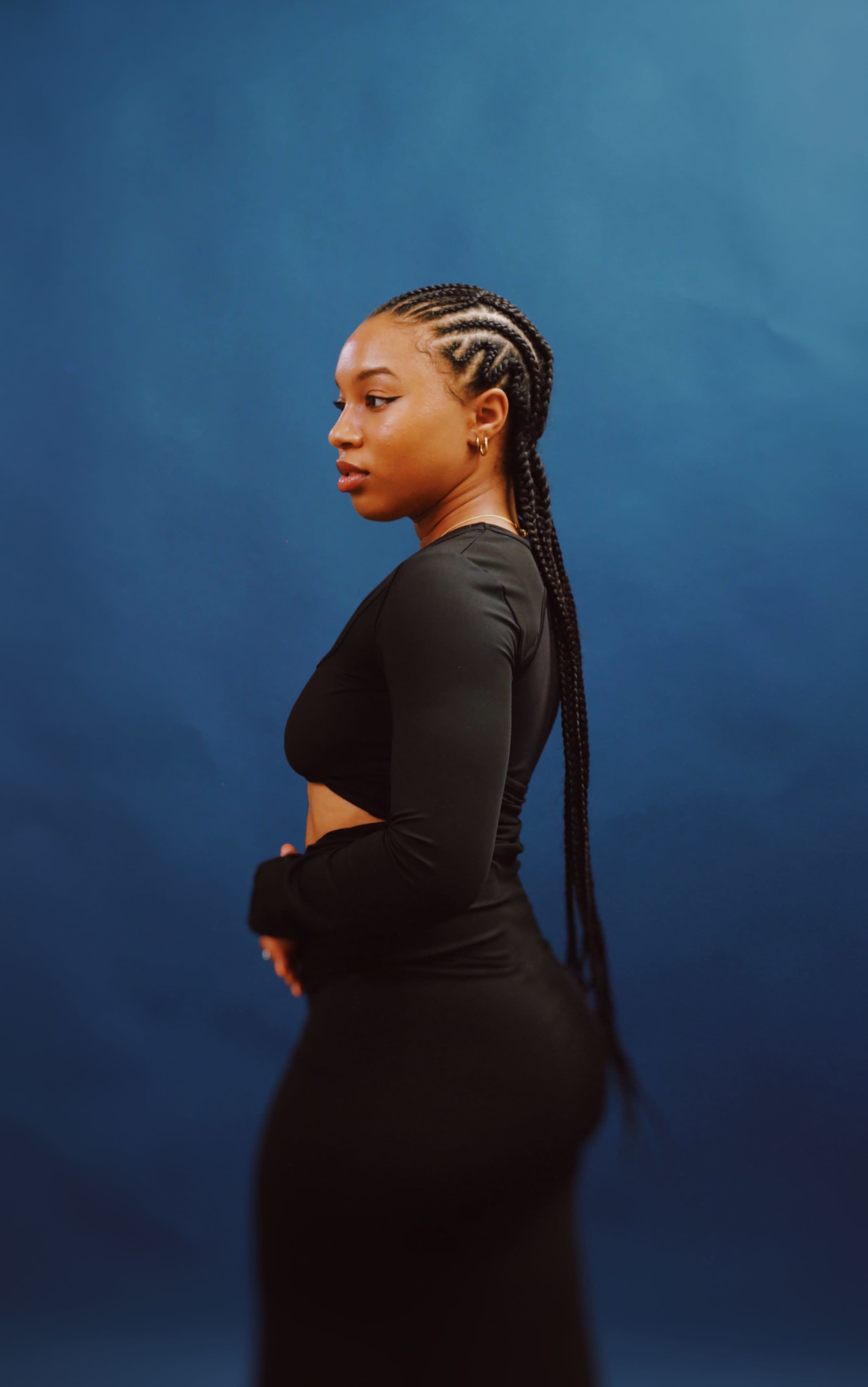 A woman with braided hair poses against a blue backdrop while wearing a fitted black top and pants.