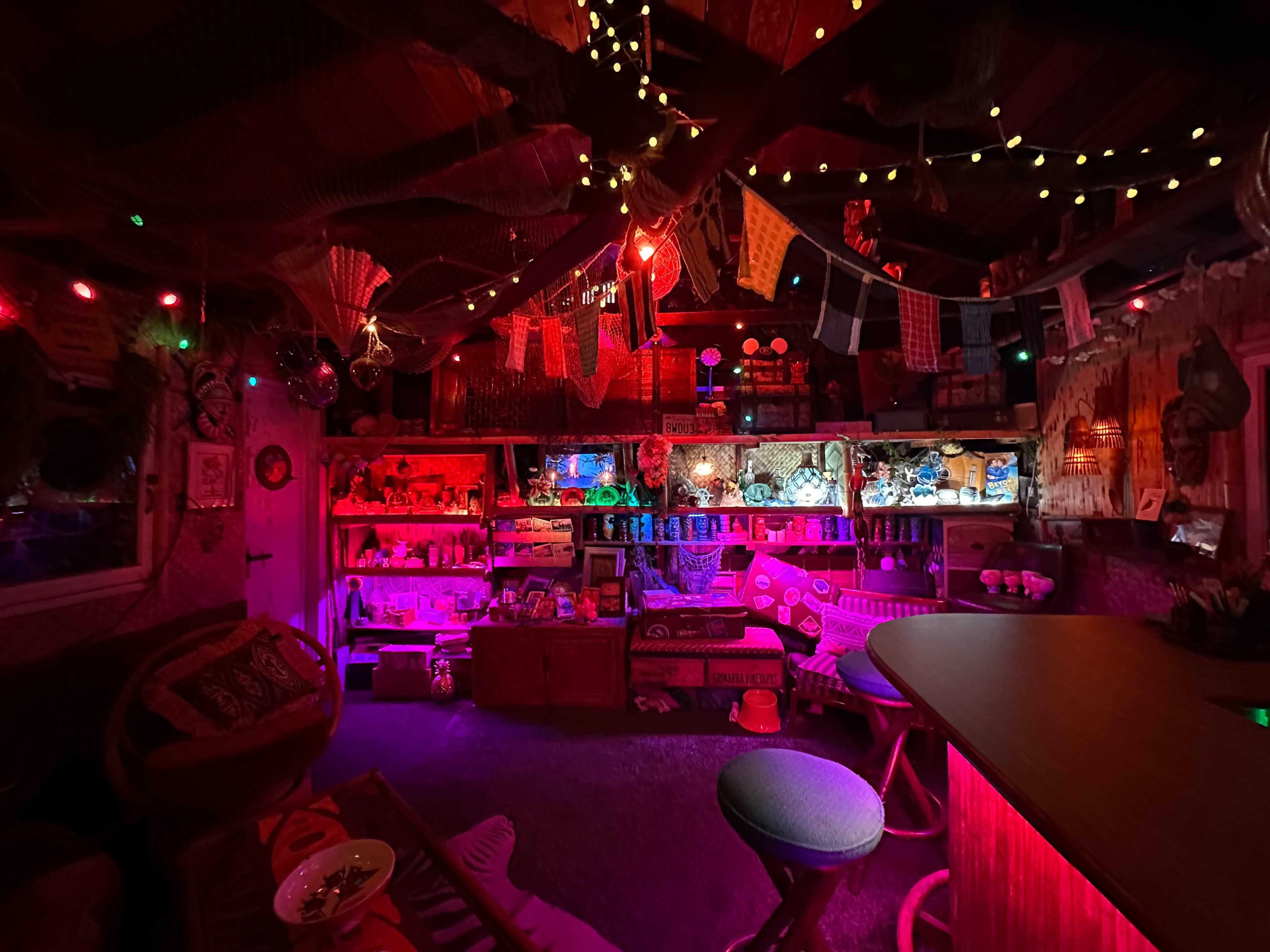 A dimly lit bar area with colorful lighting, featuring shelves filled with decorative items and a circular bar with stools.