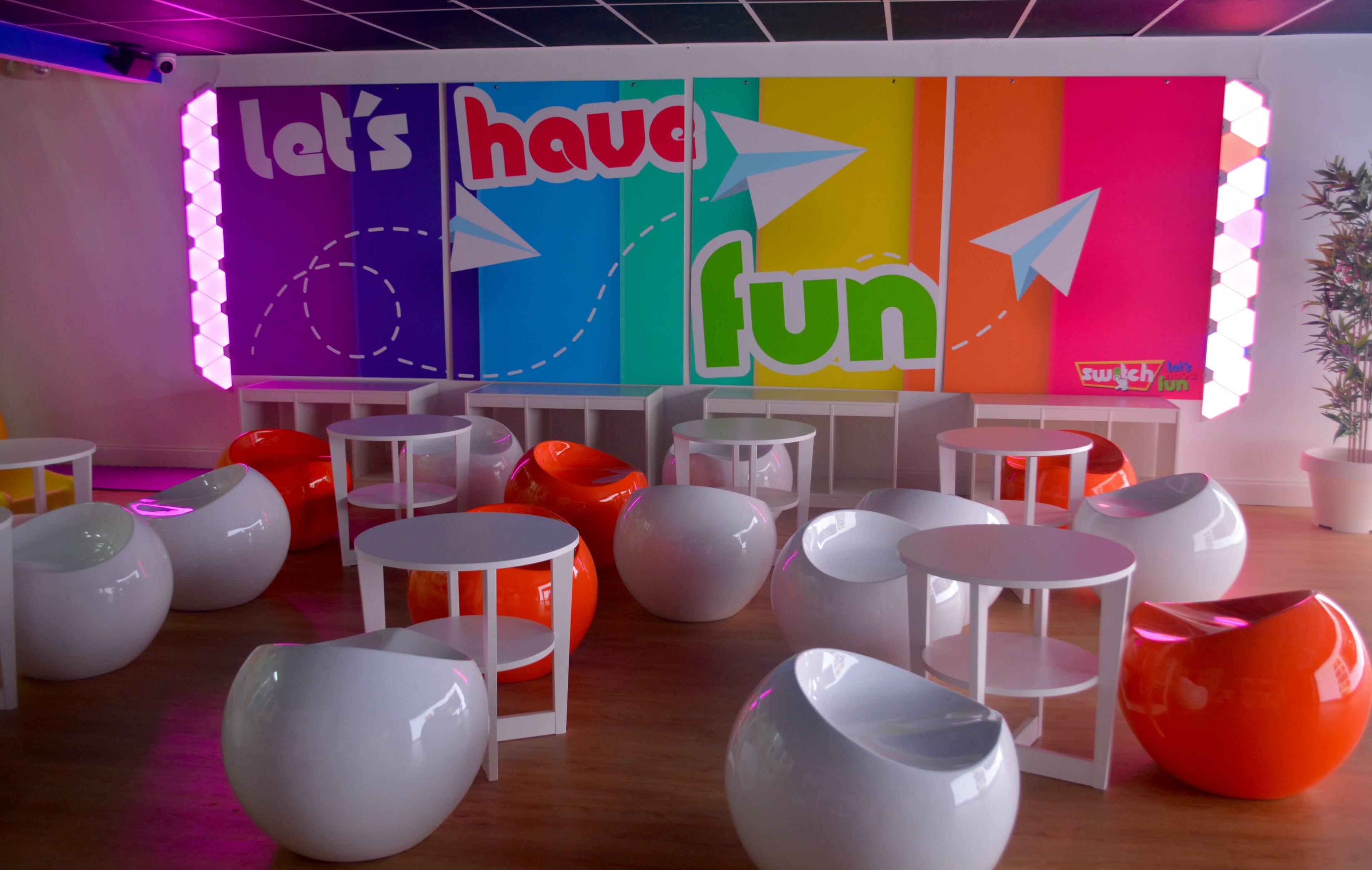 A brightly colored room features white and orange rounded chairs arranged around white tables, with a vibrant mural on the wall that reads "let's have fun."