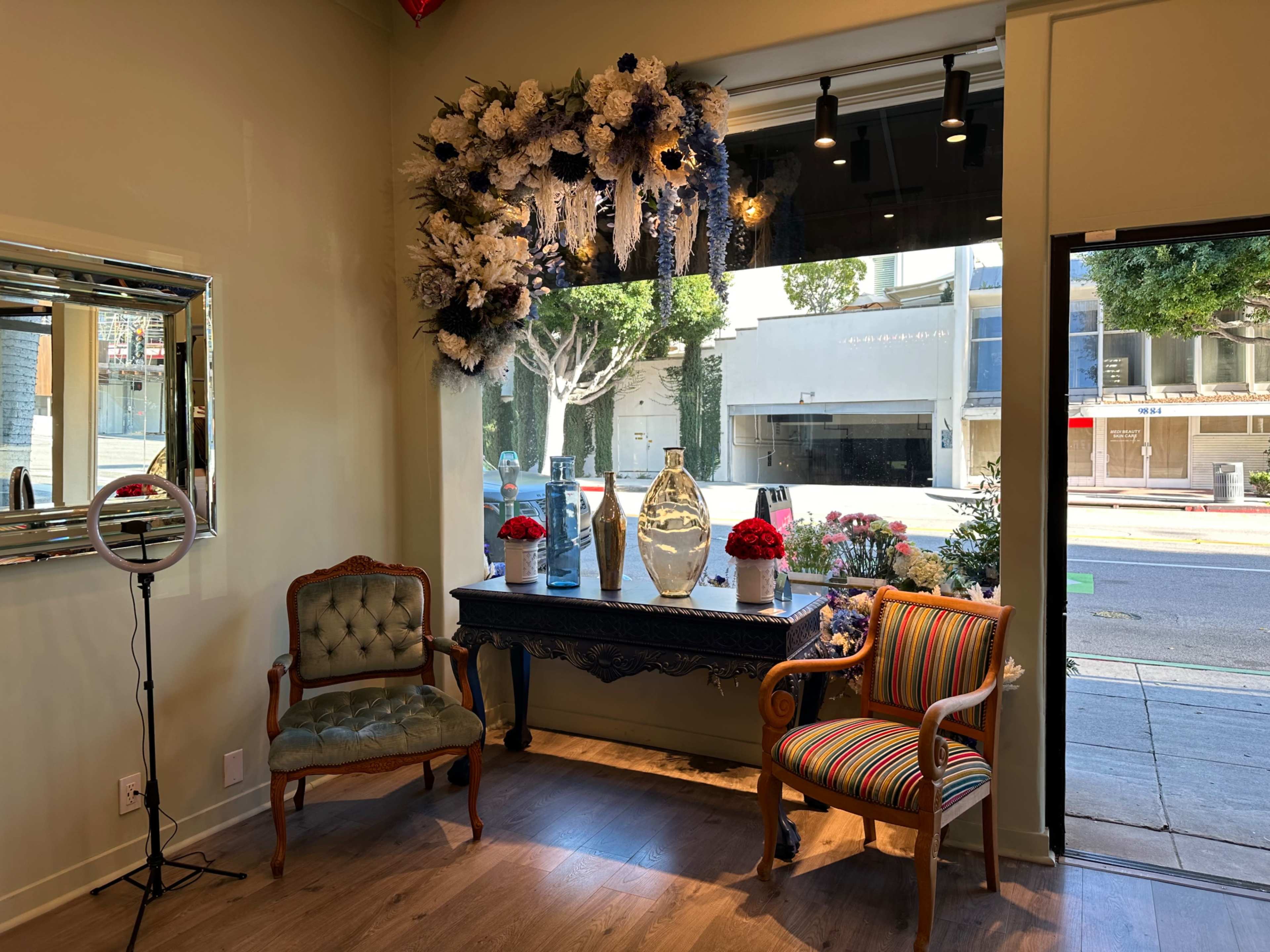 The image shows a seating area in a storefront with two chairs, a decorative table, and floral arrangements displayed in a window.