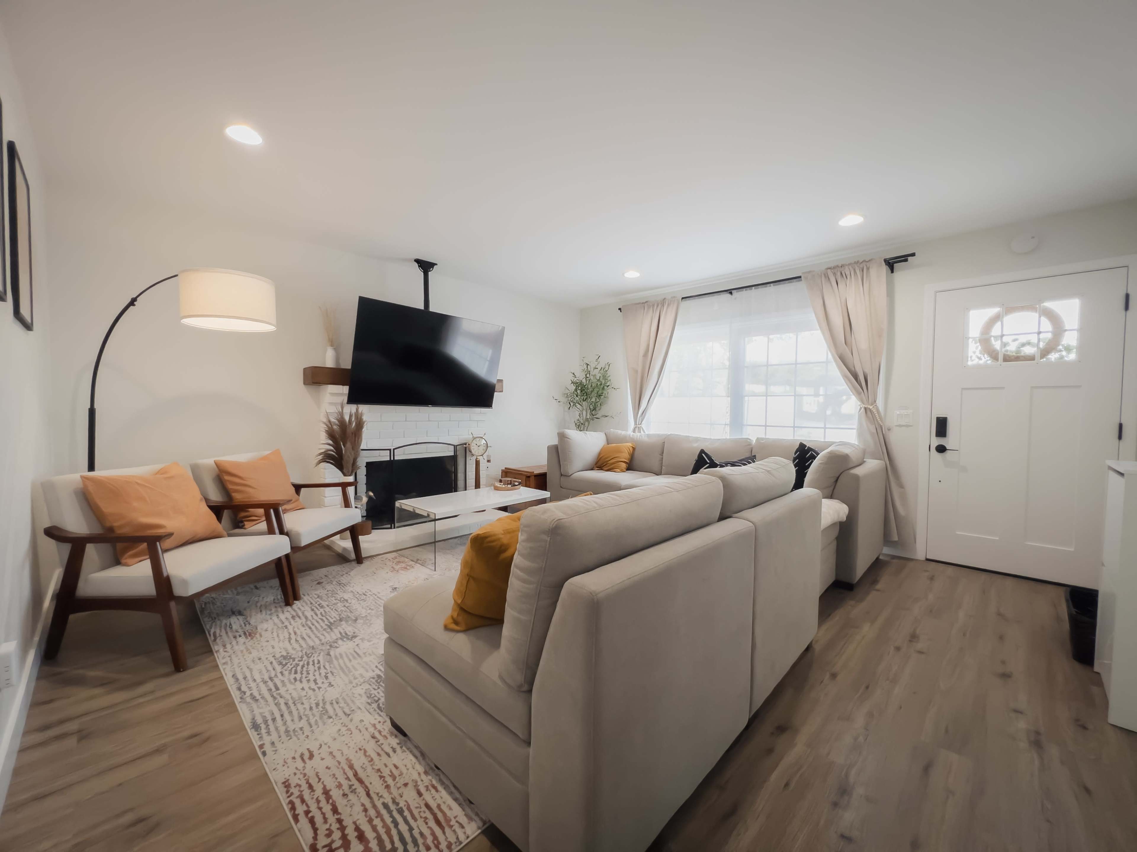 The room features a cozy living area with a large sectional sofa, two armchairs, a coffee table, a fireplace, and a mounted television, all illuminated by natural light from a nearby window.