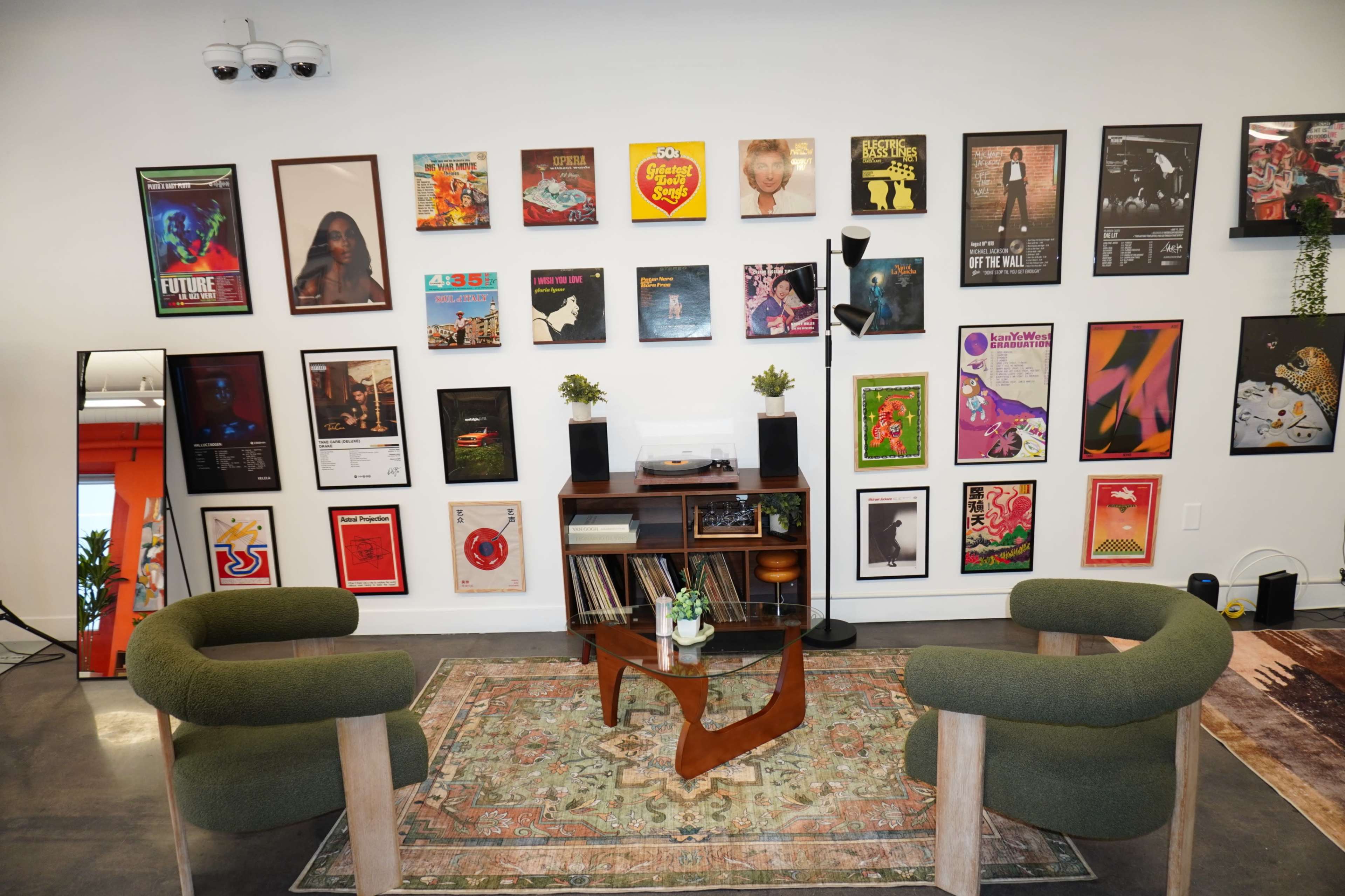 The image shows a modern living space featuring a wall adorned with various framed posters, two green armchairs, a wooden table, and decorative plants.