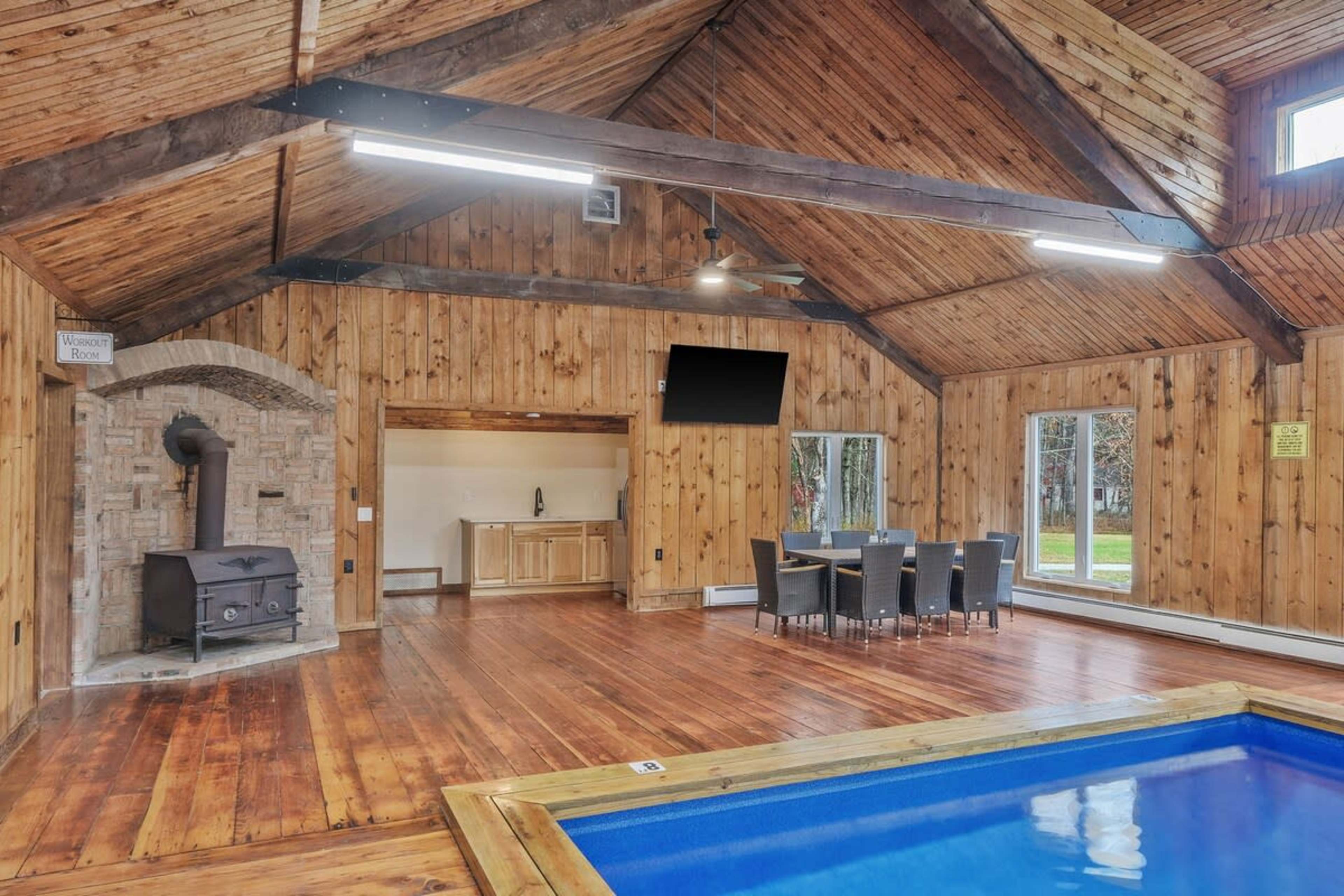 The image shows a spacious, wooden interior with a swimming pool, dining area, and a stove, illuminated by overhead lights.