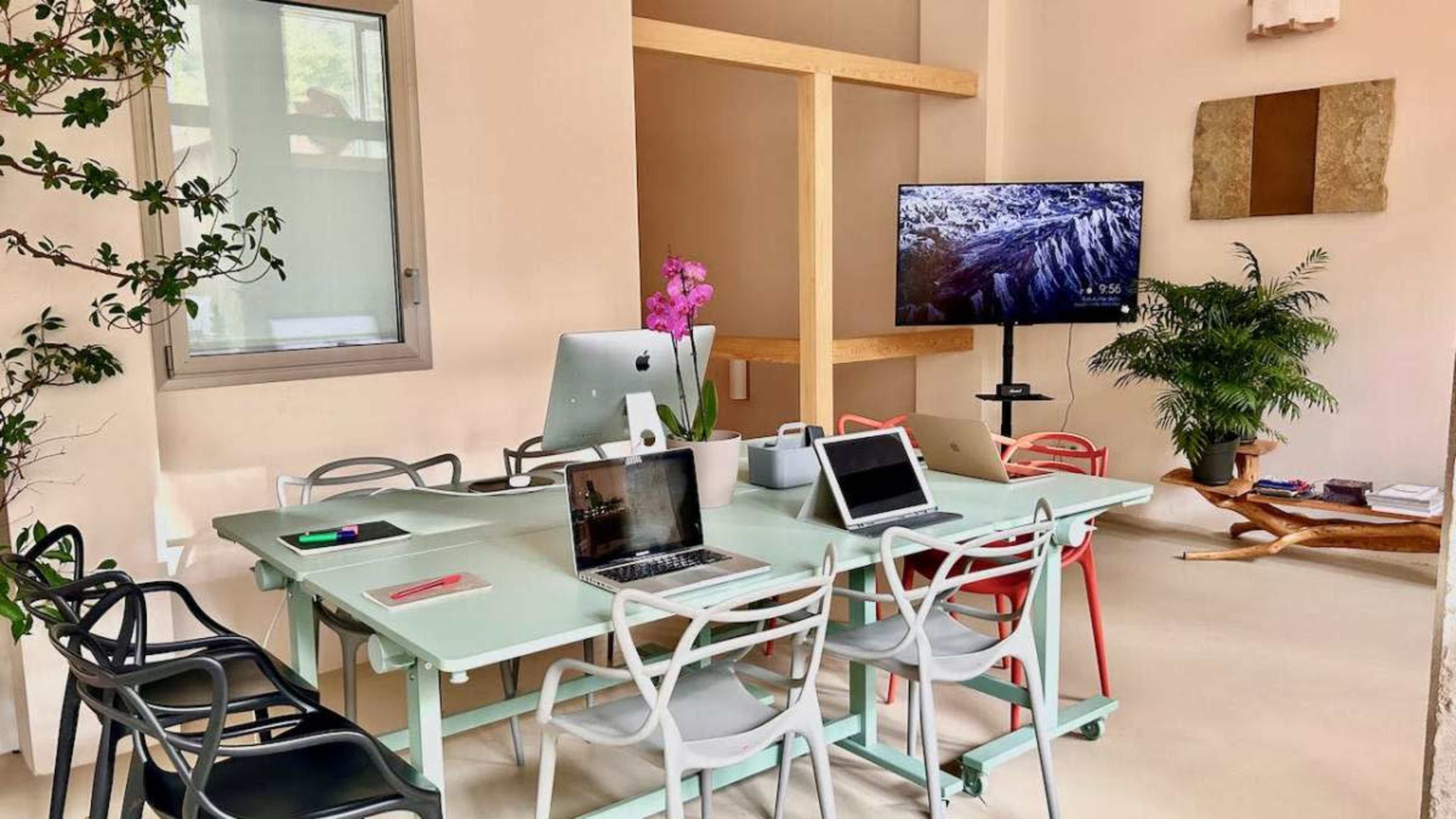 The image shows a modern meeting room featuring a large table surrounded by colorful chairs, a large television screen, and various electronic devices on the table.