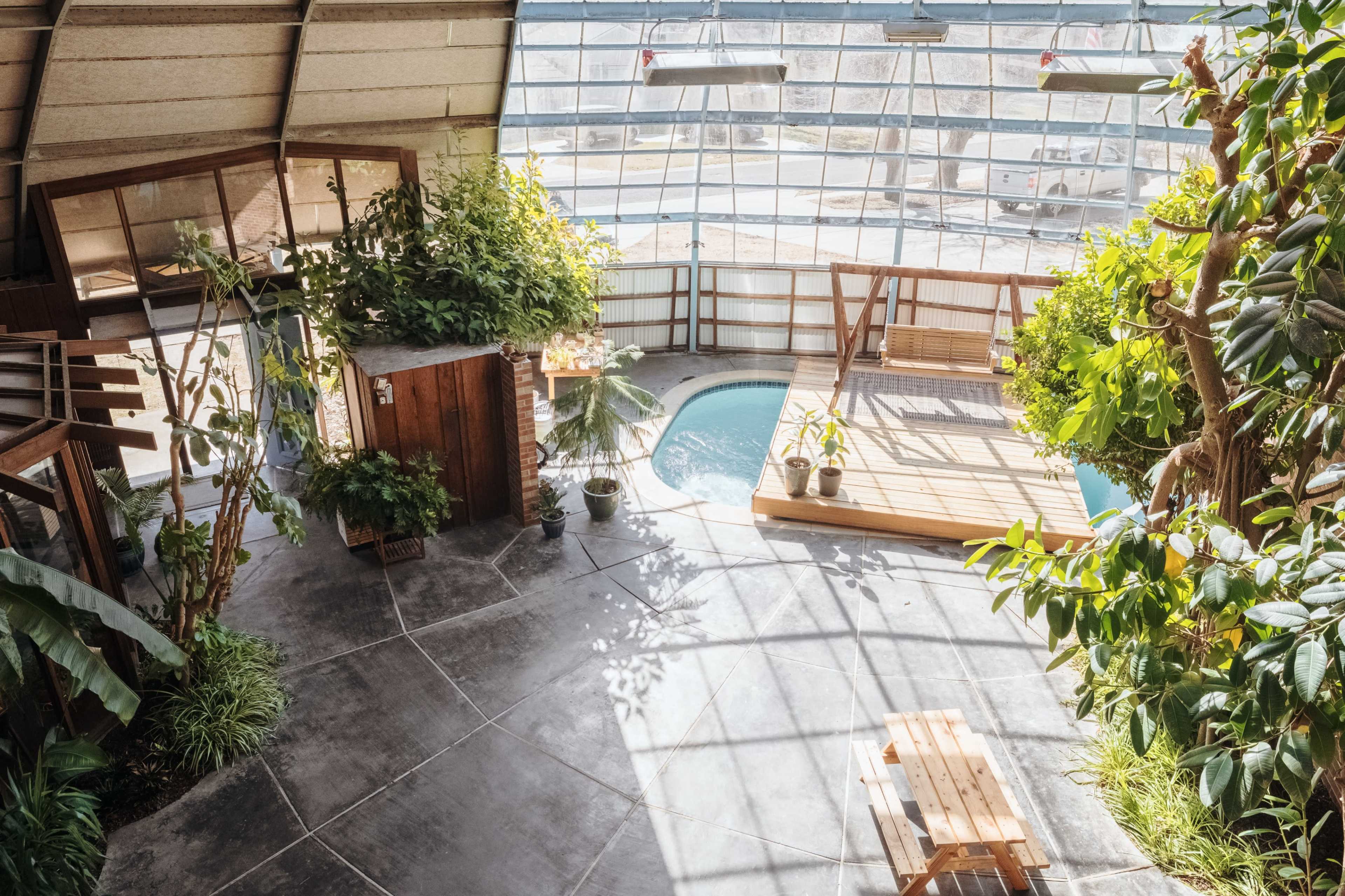 A spacious indoor area with a wooden deck, a small pool, lush greenery, and large windows allowing natural light to flood the space.