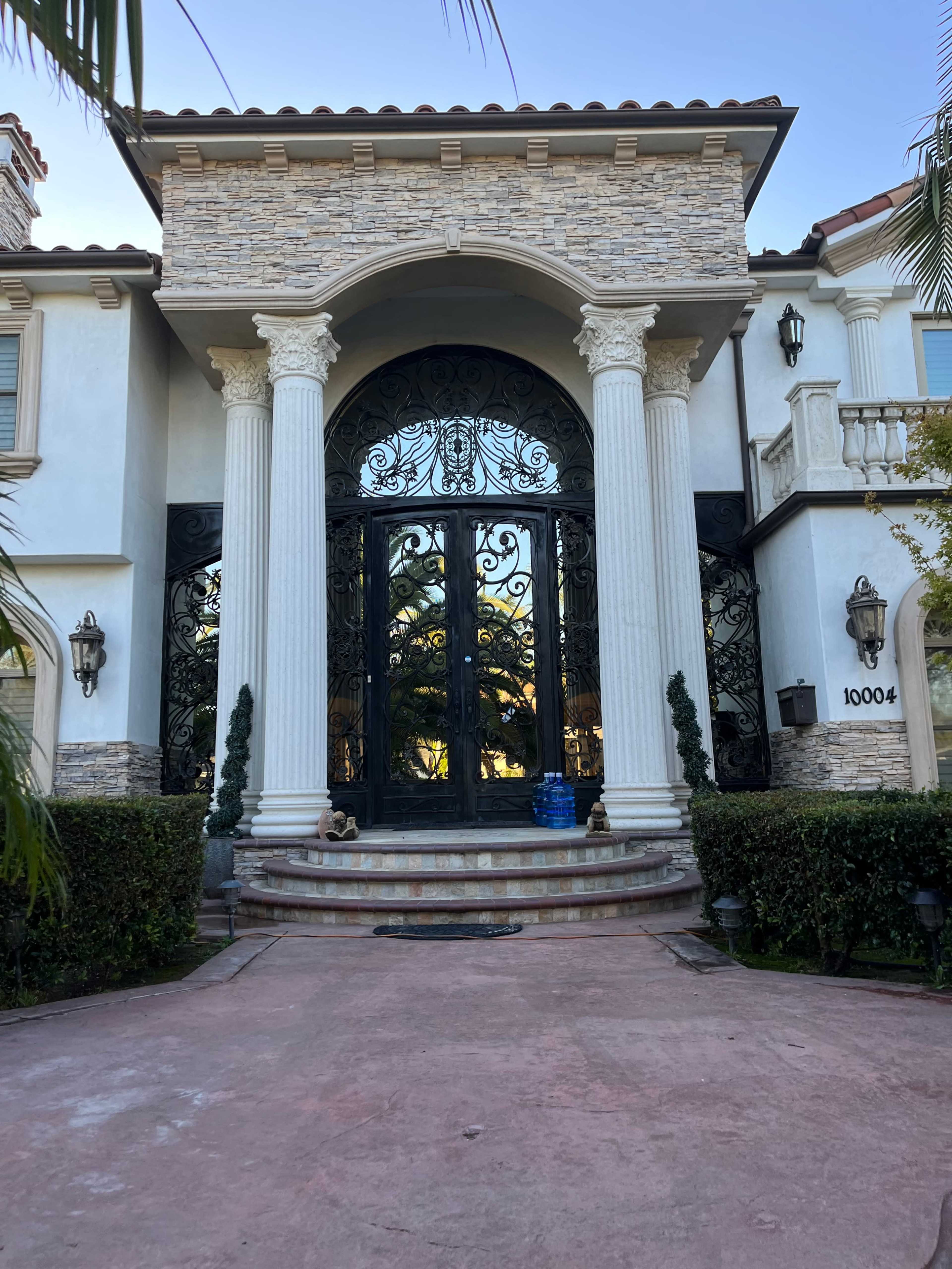A large, ornate entrance of a luxurious home with decorative columns, a grand double door, and landscaped surroundings.