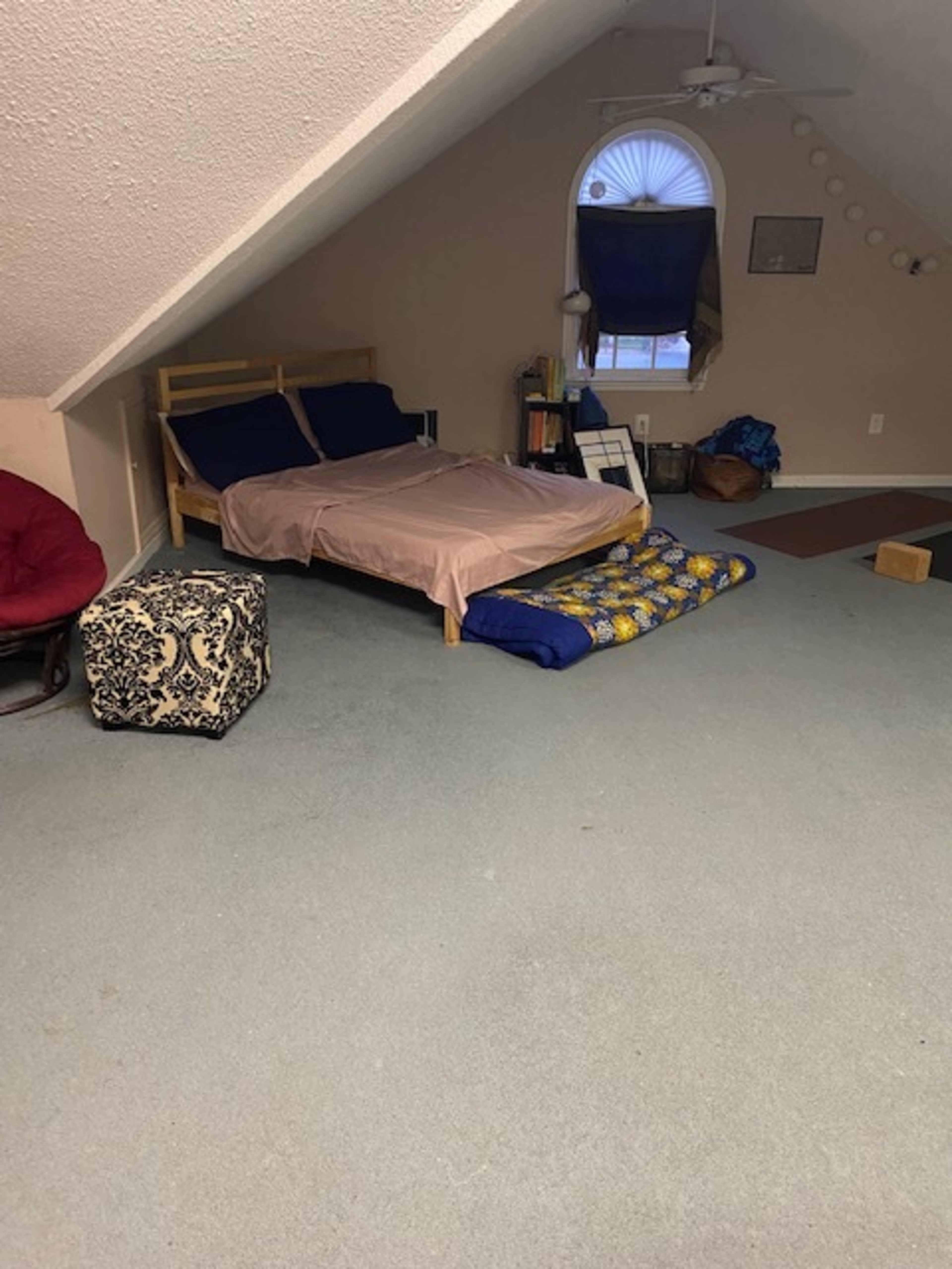 The image shows an attic bedroom with a wooden bed, a blue mat on the floor, and a small padded ottoman in the corner.