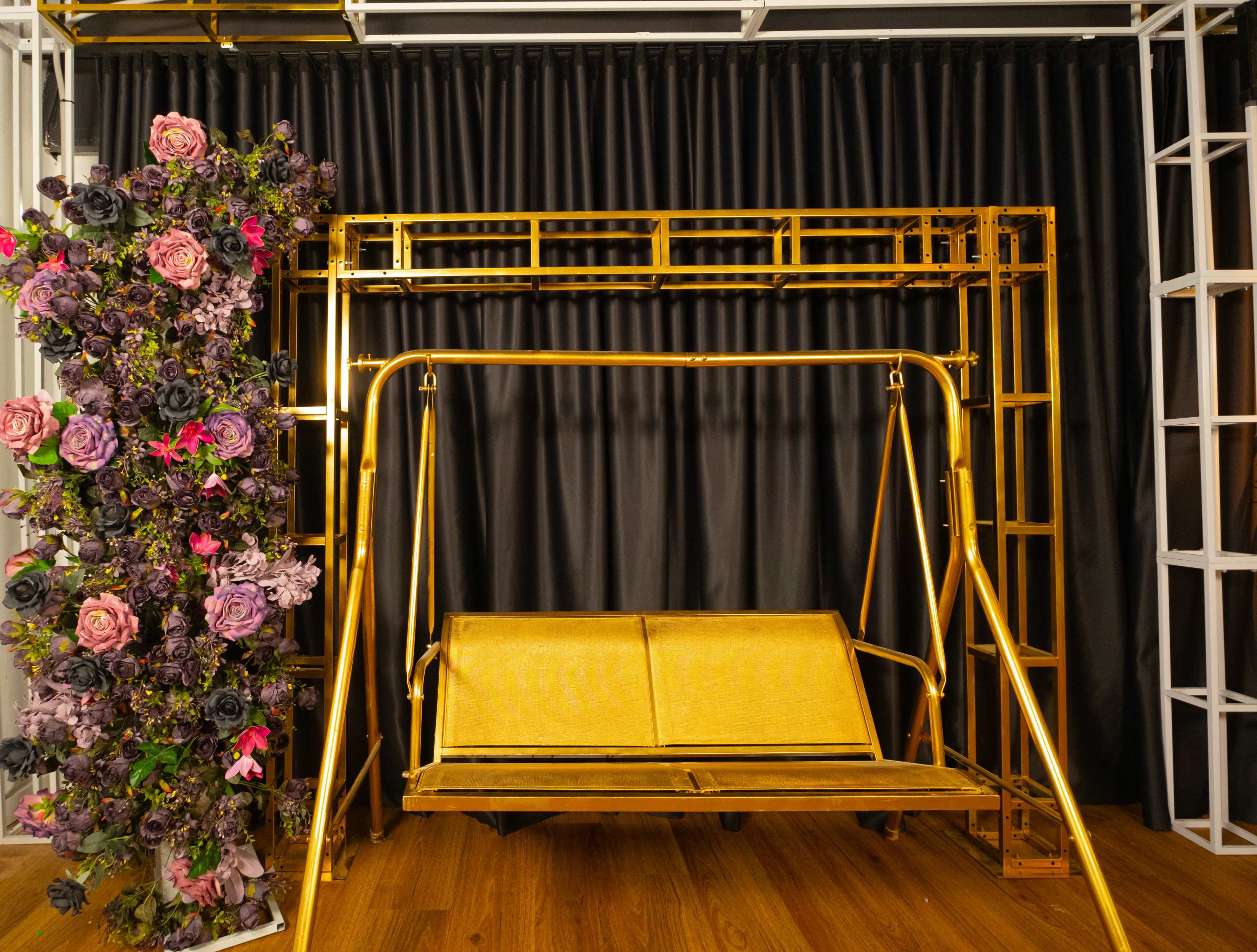 A gold-colored swinging bench is positioned in front of a black curtain and is flanked by a floral arrangement adorned with purple and pink flowers.