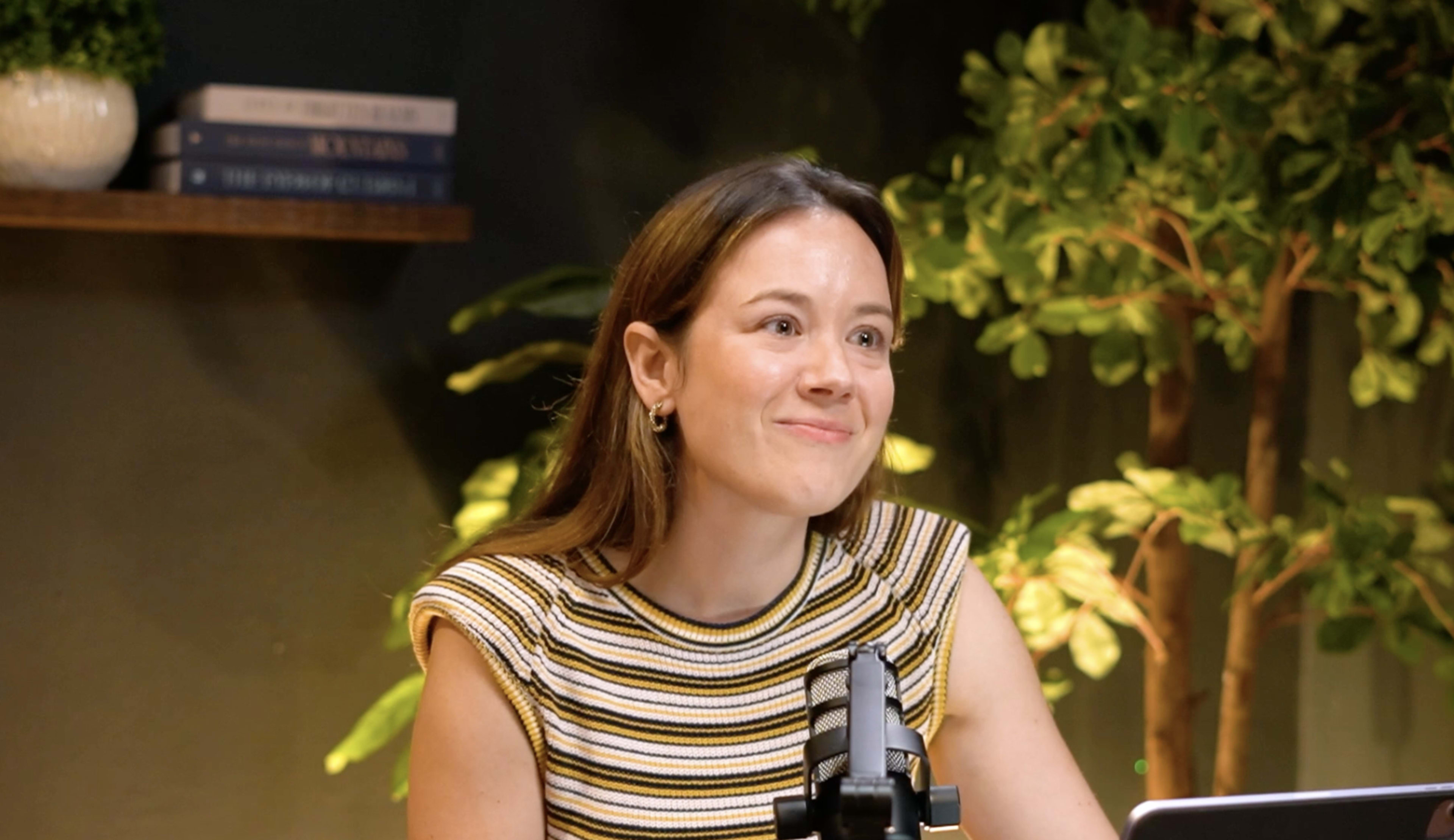 A woman in a striped shirt sits at a desk with a microphone in front of her, smiling amid green plants and books on a shelf in the background.