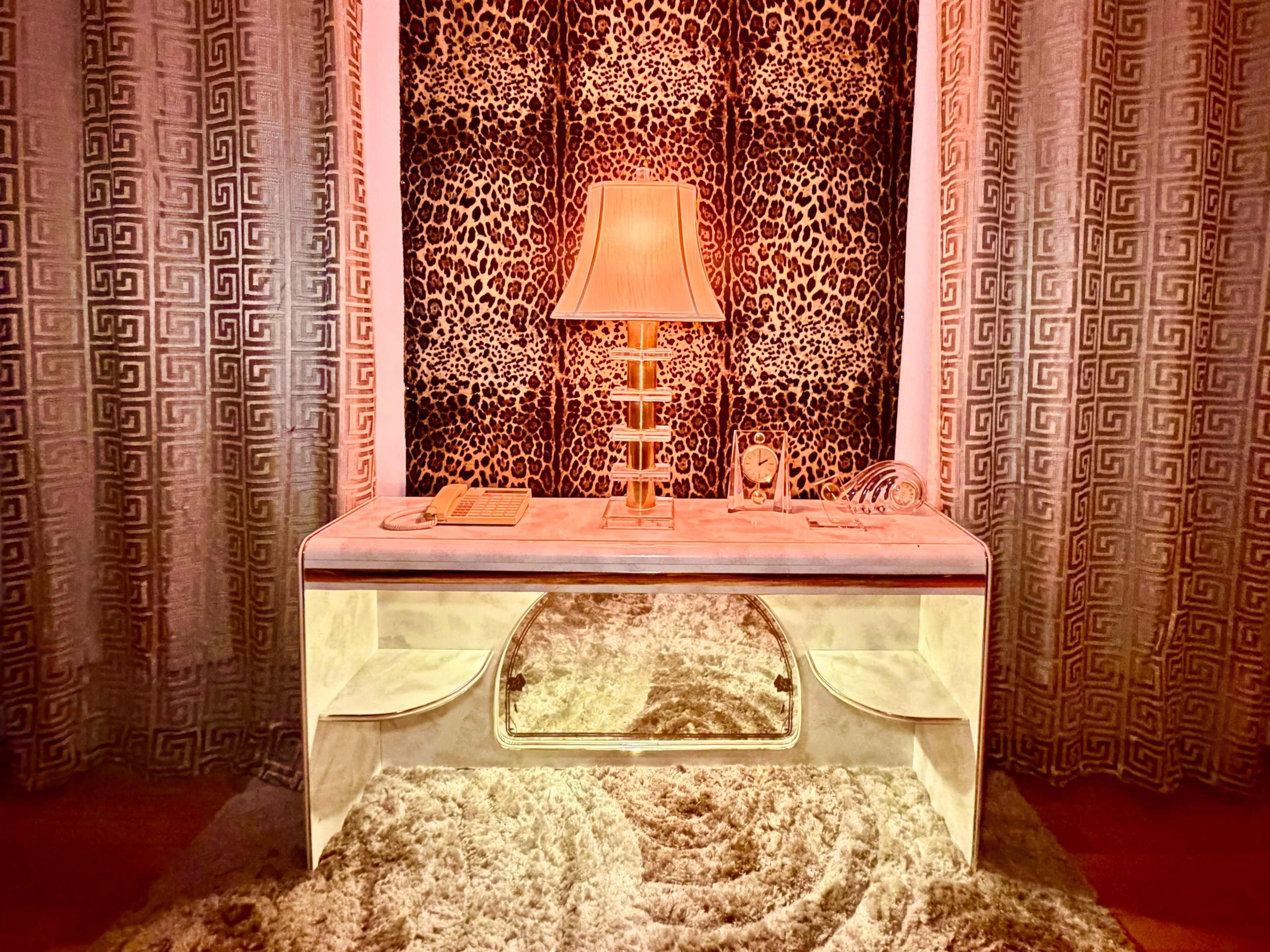 The image features a stylish desk with a mirror front, a gold lamp, and decorative items, set against a backdrop of leopard-print curtains and a plush area rug.