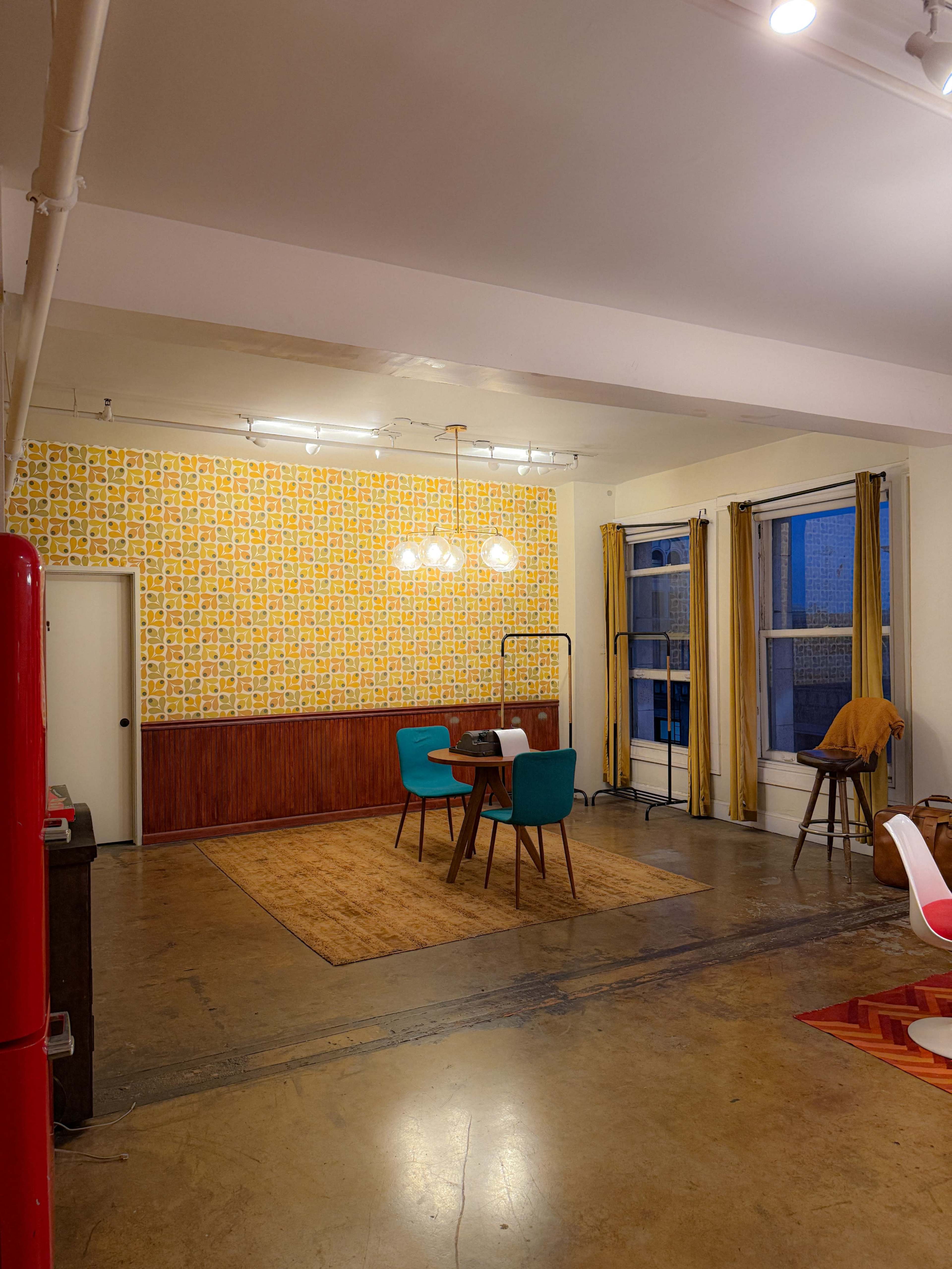The image shows a spacious room with a dining area featuring a round table and four chairs, a patterned yellow wallpaper, and large windows behind light-colored curtains.