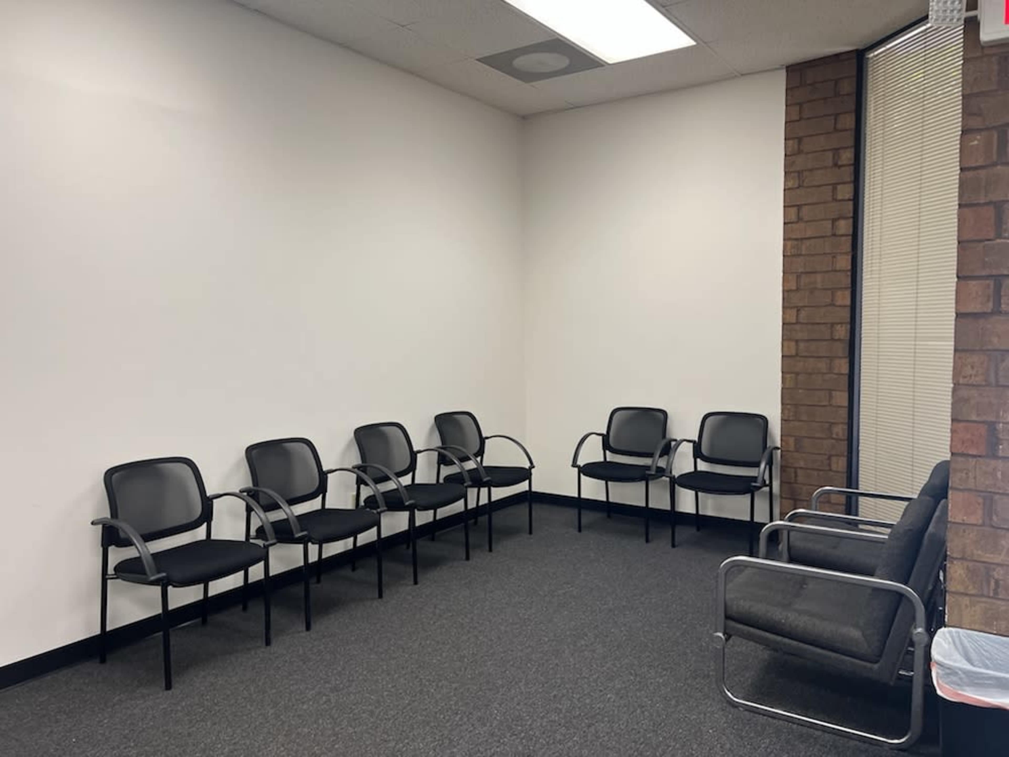 The image shows a waiting room with several black chairs arranged against a plain white wall.