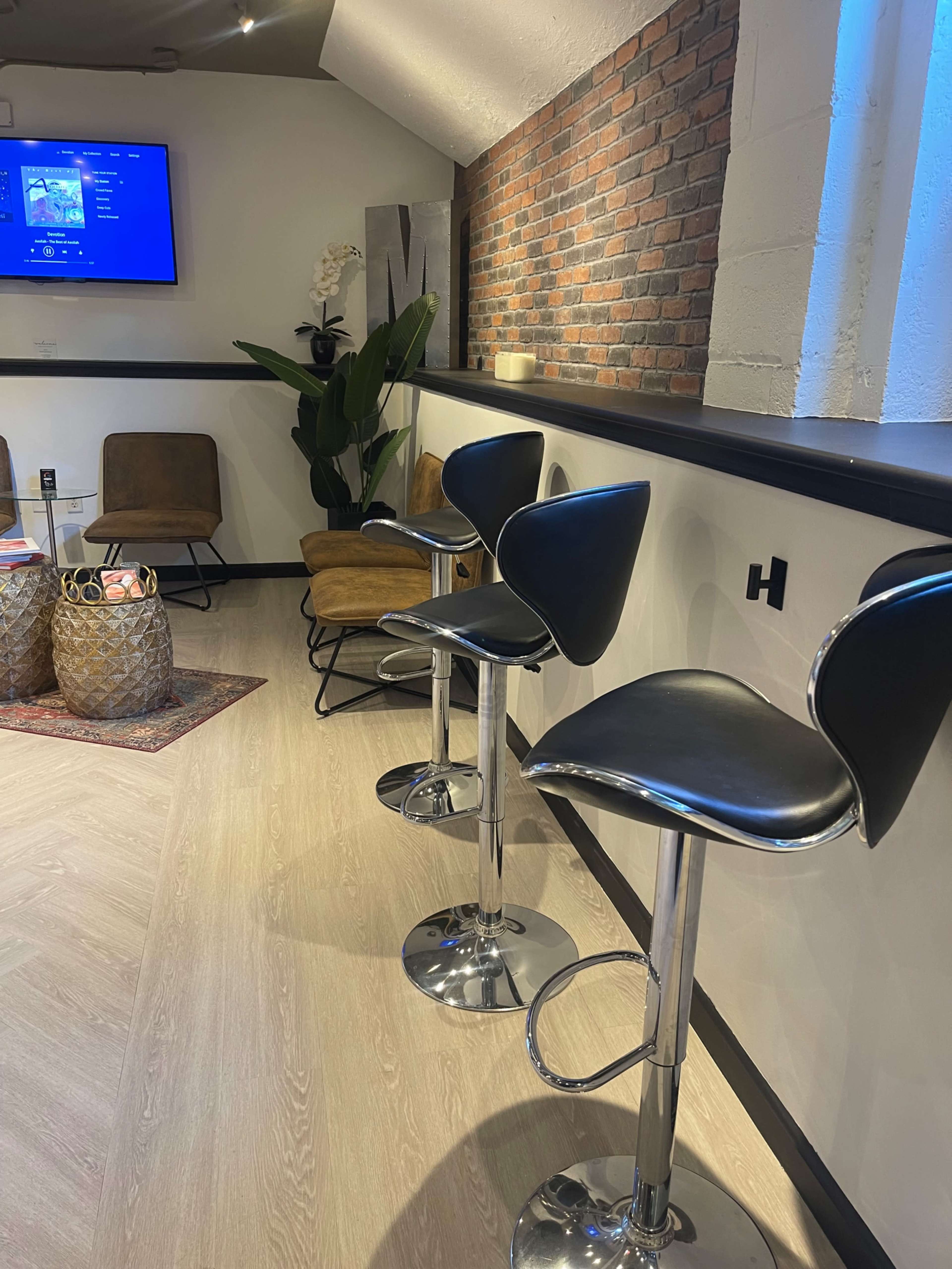 The image shows a modern interior space with bar stools arranged along a counter, a brick wall, and a plant in the corner.